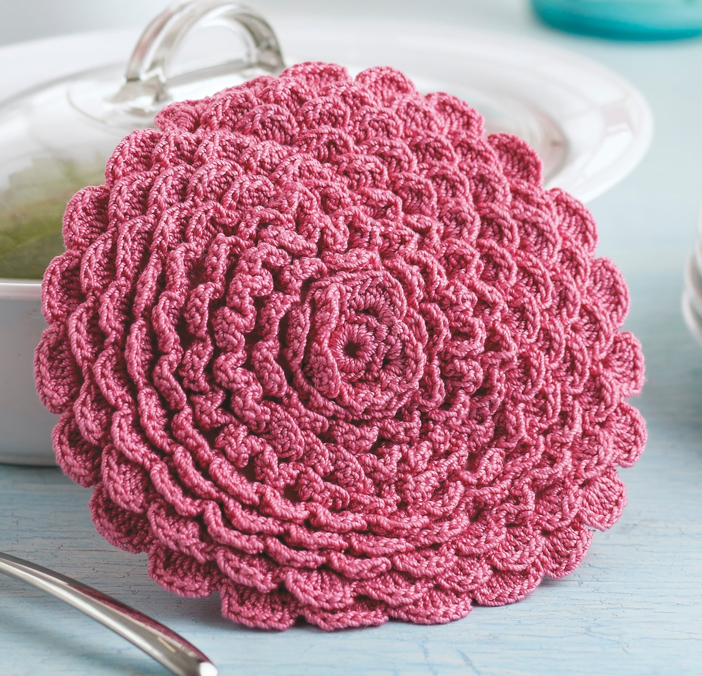 The Potholders & Dishcloths Digital Download from Leisure Arts features a round pink crocheted potholder resembling a blooming rose—layered spiral petals and scalloped edge—ideal for unique crochet gift ideas.