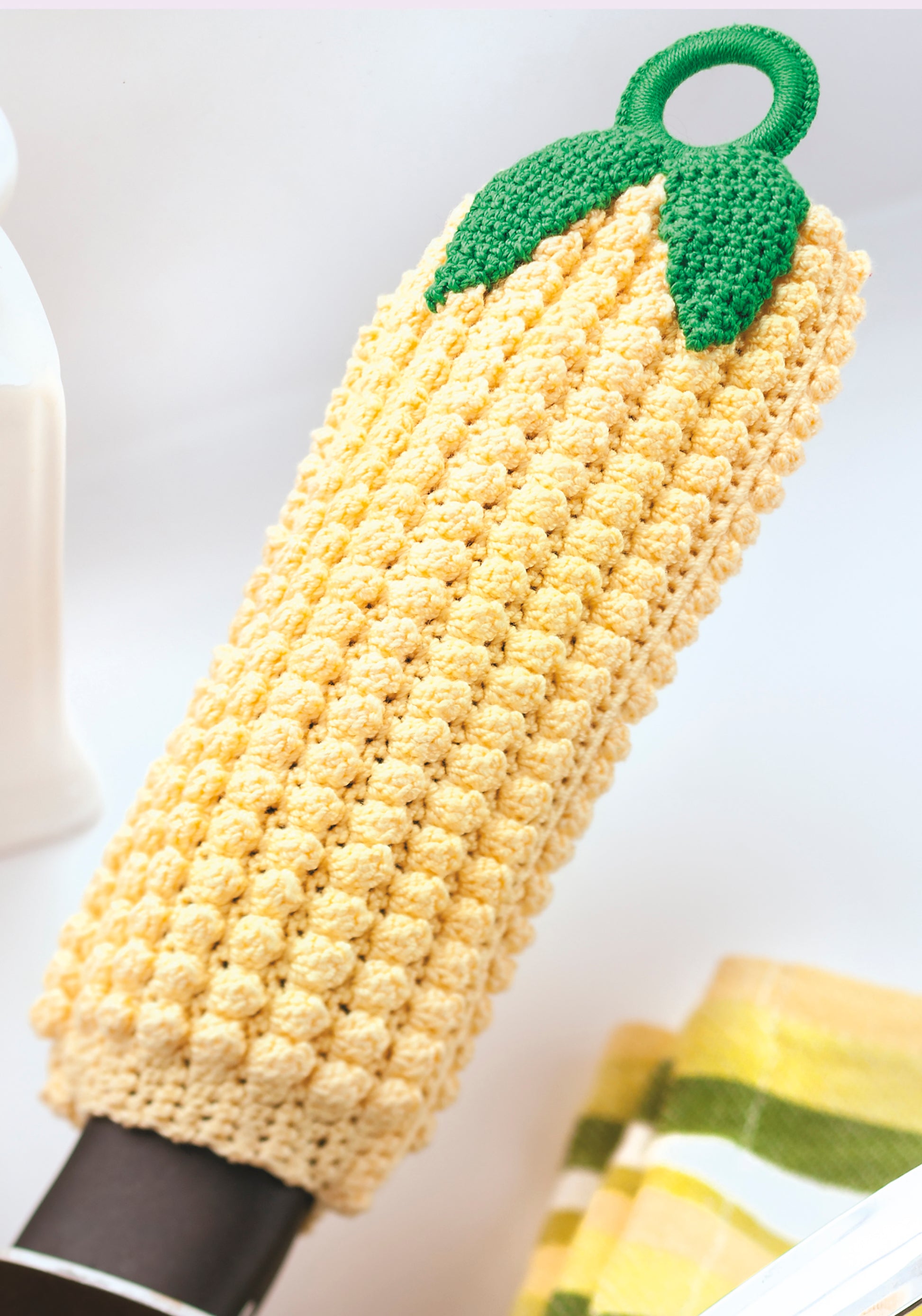 A yellow, textured crochet bottle cover resembling a corn cob sits by a striped towel, made using Leisure Arts' Potholders & Dishcloths Digital Download—a great source for unique crochet gift ideas.