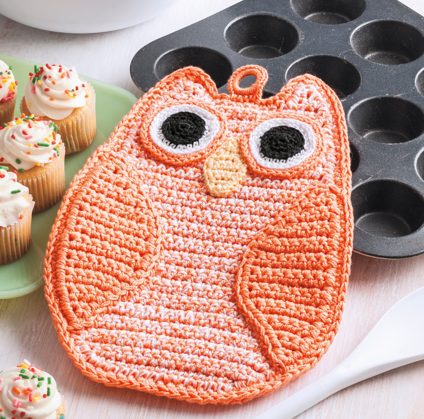 A crocheted orange owl potholder from Leisure Arts' Potholders & Dishcloths Digital Download sits next to a muffin tin, frosted mini cupcakes with sprinkles, and a wooden spoon on a white surface.