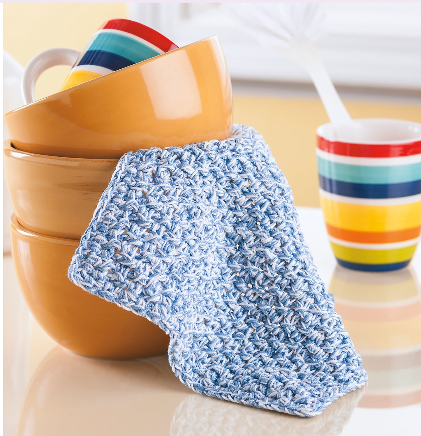 A crochet blue cotton dishcloth from Leisure Arts’ Potholders & Dishcloths Digital Download rests beside golden yellow bowls and a colorful striped mug—an inspiring choice for unique crochet gift ideas.