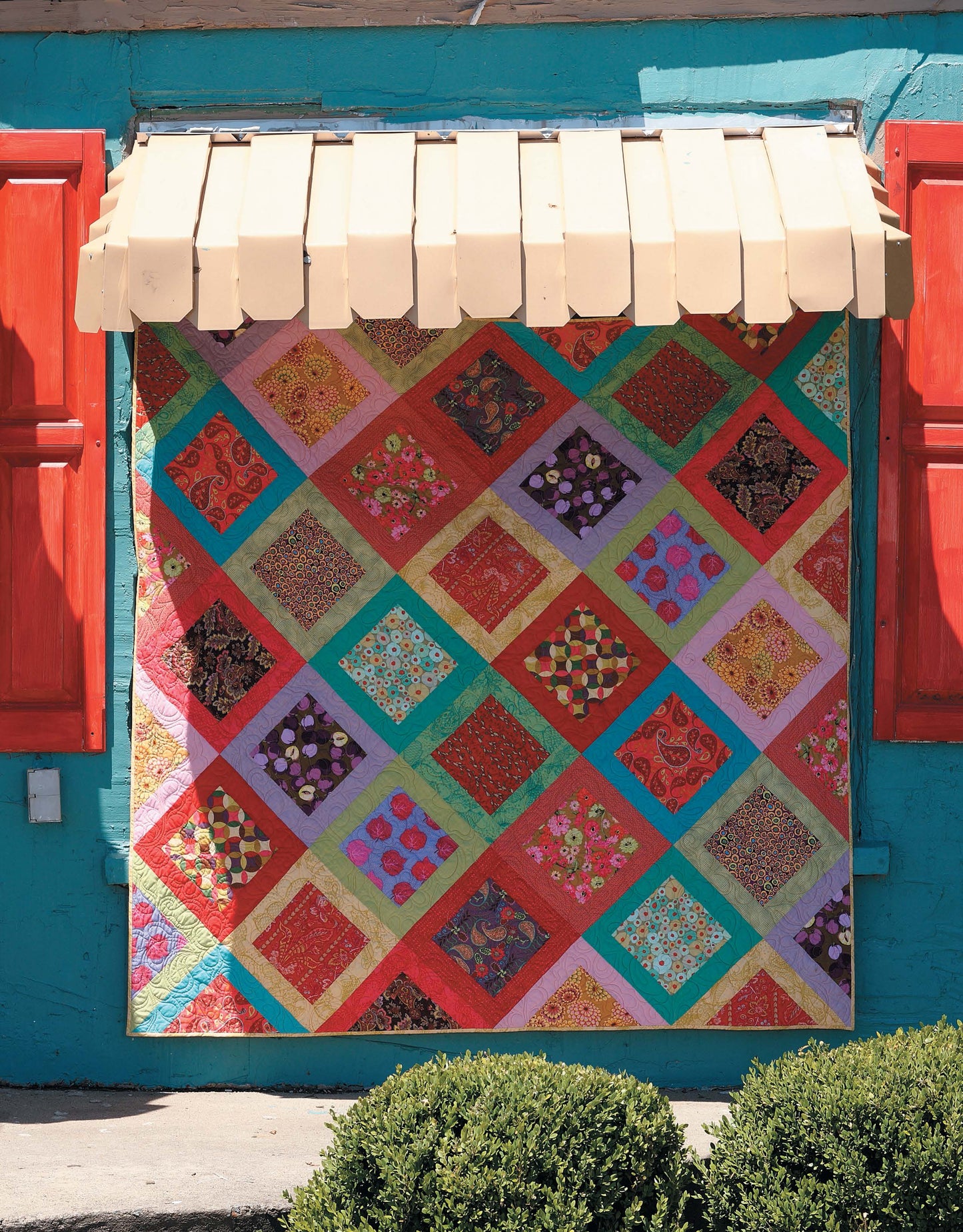 The Big Block Quilts by Leisure Arts, featuring vibrant diamond patterns in reds, greens, purples, and yellows, hangs on a blue wall with red shutters and green shrubs below—ideal for quick quilting projects.