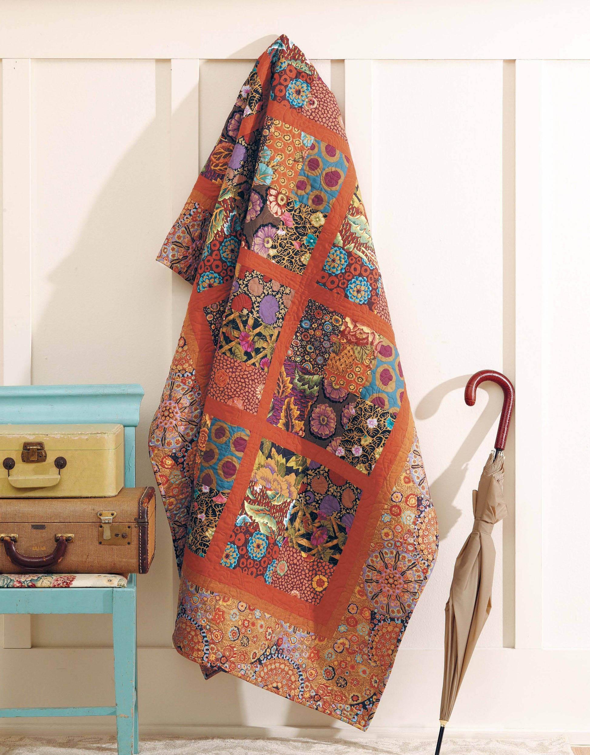 The "Big Block Quilts" by Leisure Arts, featuring vibrant patterns, hangs on a white paneled wall above a beige umbrella and stacked vintage suitcases resting on a blue stool.