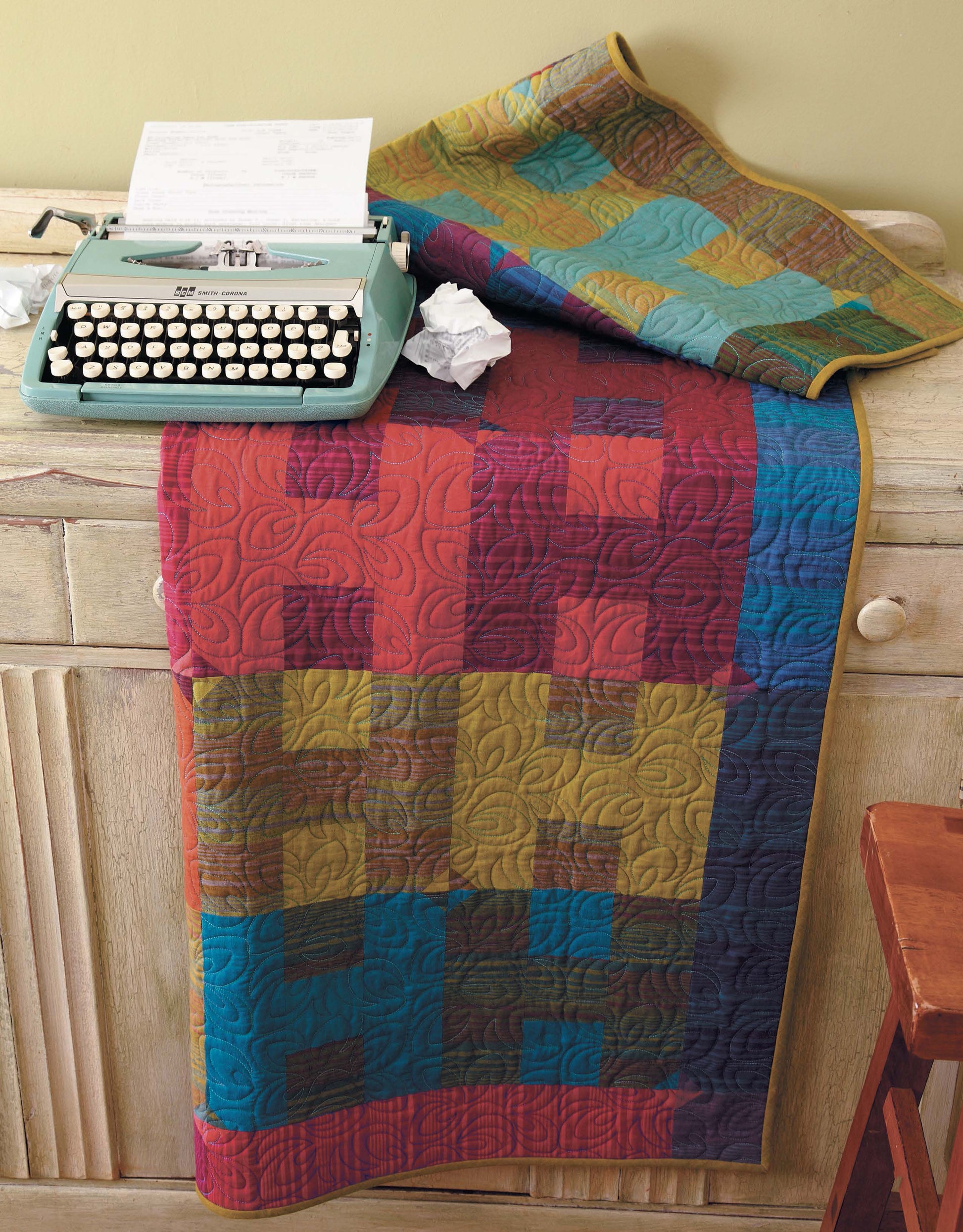The Big Block Quilts by Leisure Arts, ideal for fast quilting projects, is draped over a rustic cabinet. On top sit a vintage typewriter and crumpled paper, with part of a wooden chair visible beside the cabinet.