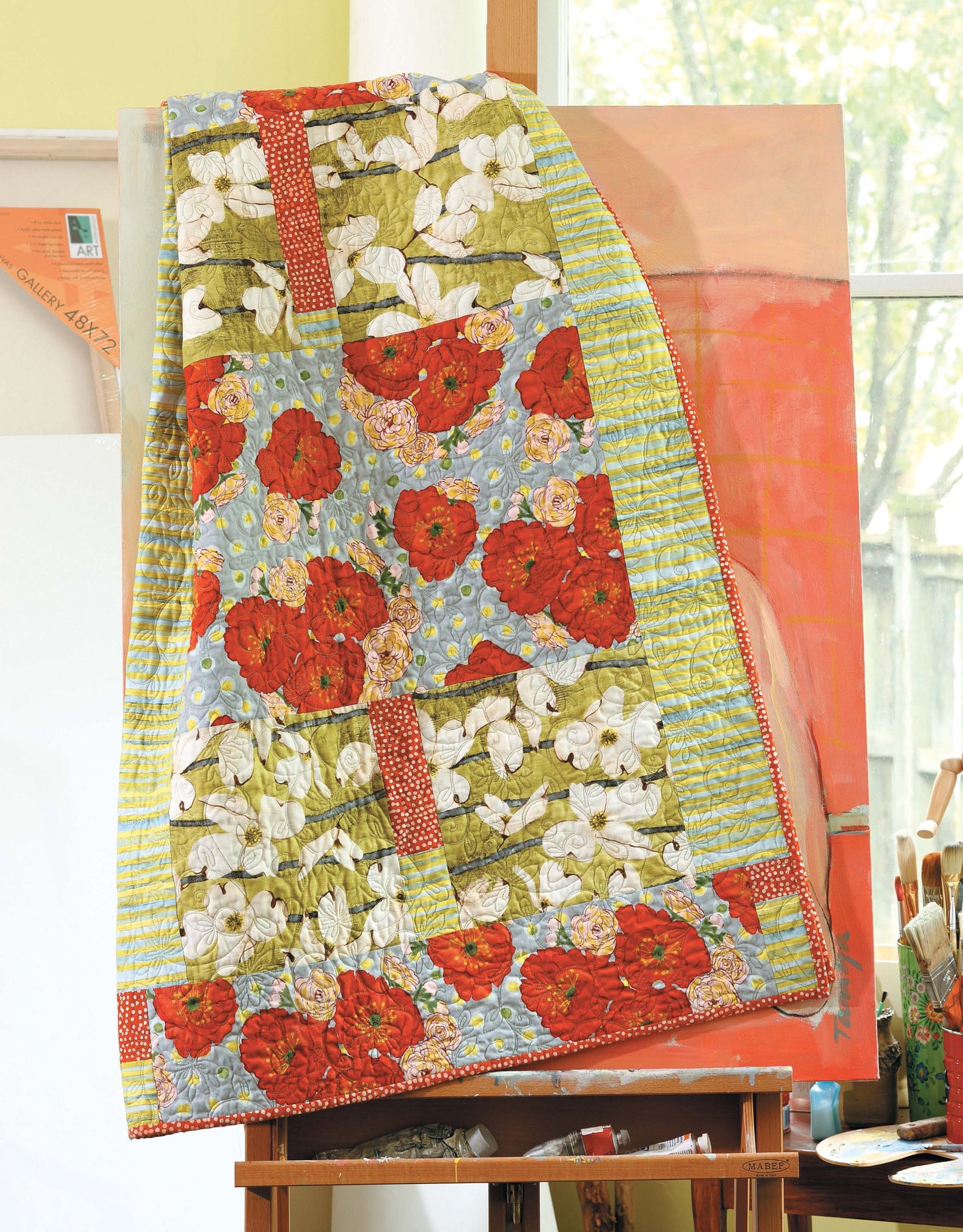 The Big Block Quilts by Leisure Arts, featuring floral patterns and vibrant red, white, blue, and green sections—ideal for fat-quarter designs—is draped over an easel in a sunlit room with painting supplies nearby.