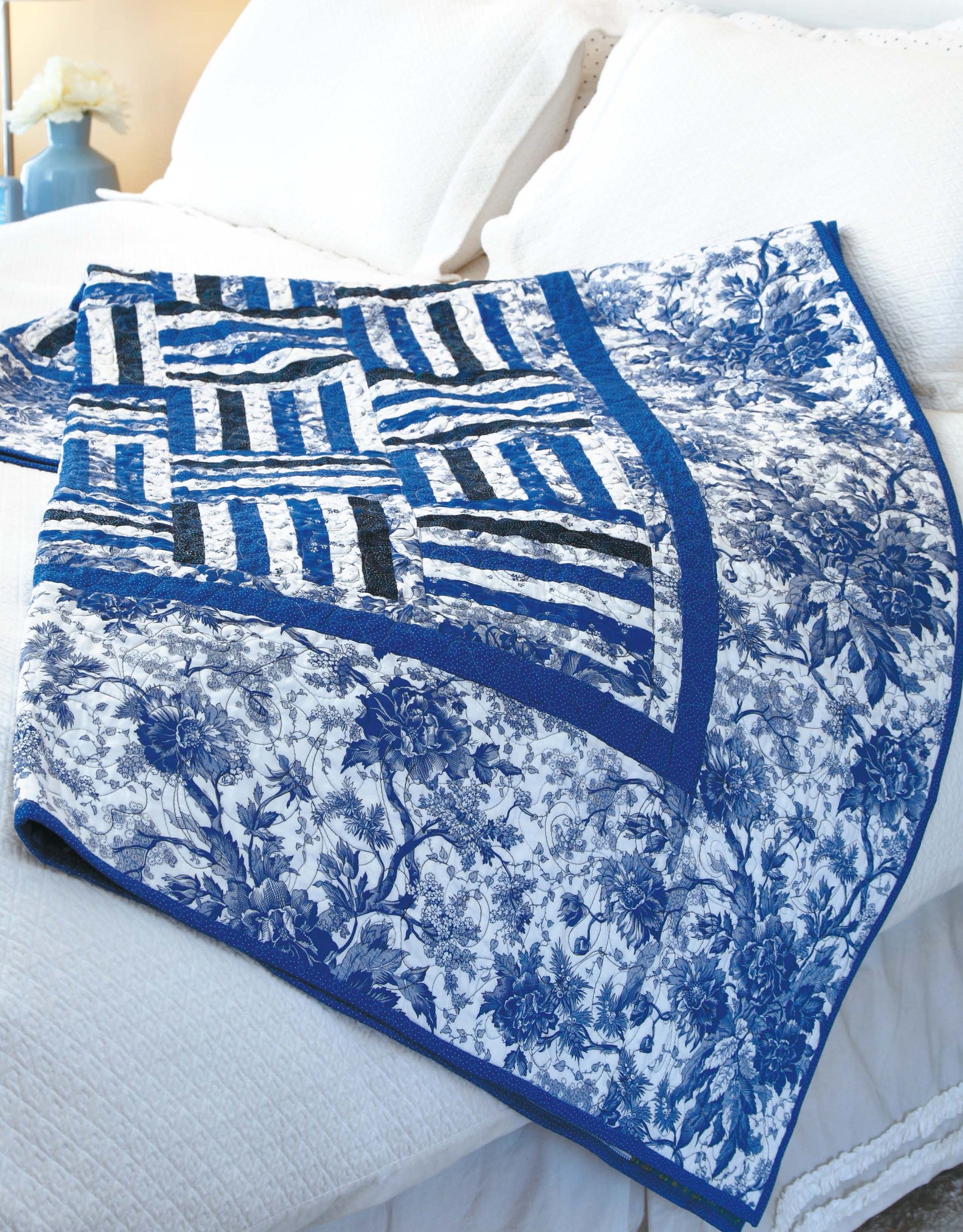 The "Big Block Quilts" by Leisure Arts, featuring blue and white floral and striped patterns, is draped over a white bed with pillows in a softly lit bedroom—ideal for fat-quarter friendly quilt designs.