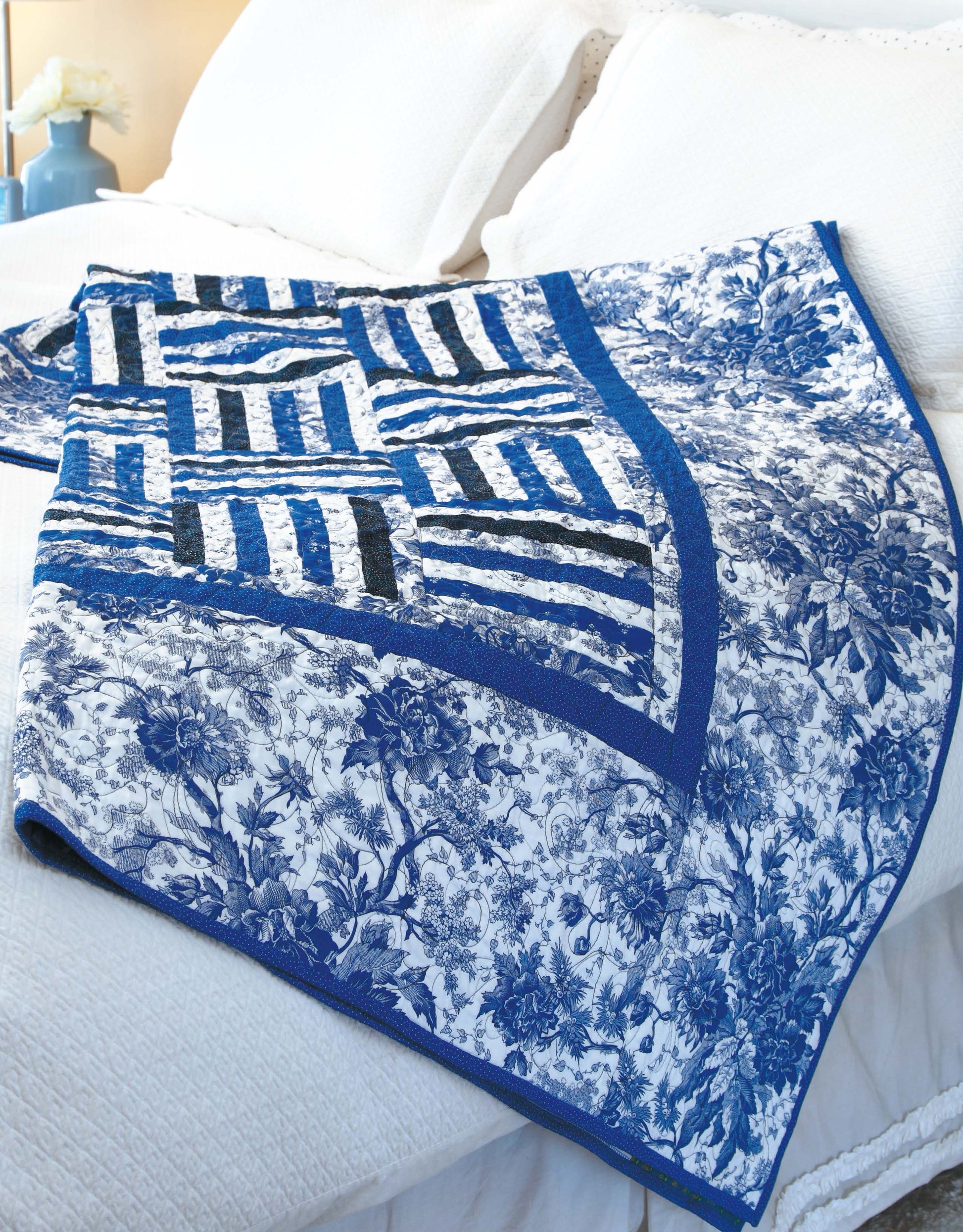The "Big Block Quilts" by Leisure Arts, featuring blue and white floral and striped patterns, is draped over a white bed with pillows in a softly lit bedroom—ideal for fat-quarter friendly quilt designs.