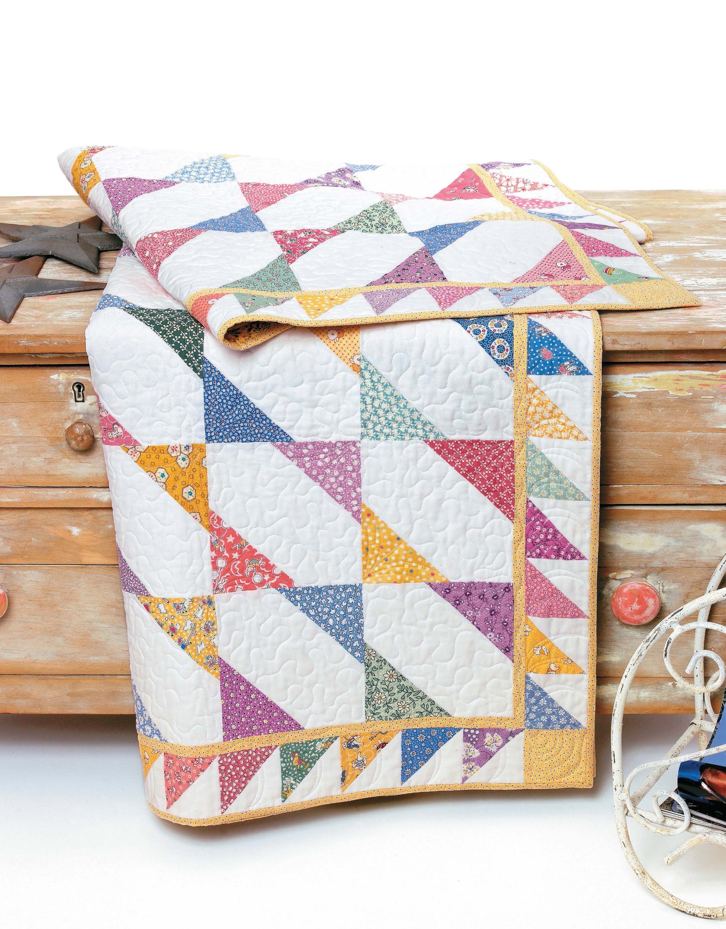 The Big Block Quilts by Leisure Arts, featuring vibrant triangle patterns in pastel prints, is draped over a rustic wooden dresser with red knobs. A white cord and metal object are partially visible on the right.