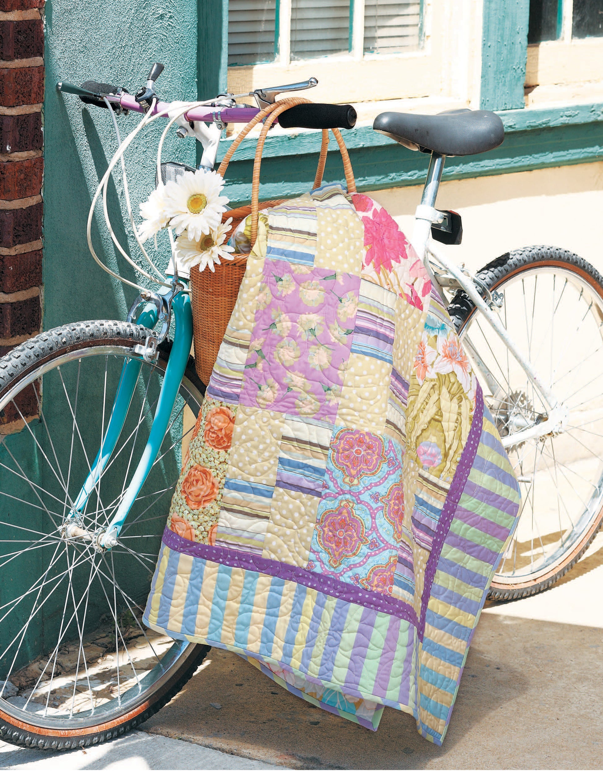 A white bicycle with a wicker basket of daisies is parked against a wall. The handlebars are draped with the Leisure Arts "Quilts for Kids—From Crib to College," inspired by baby quilt patterns and fat quarter quilts.