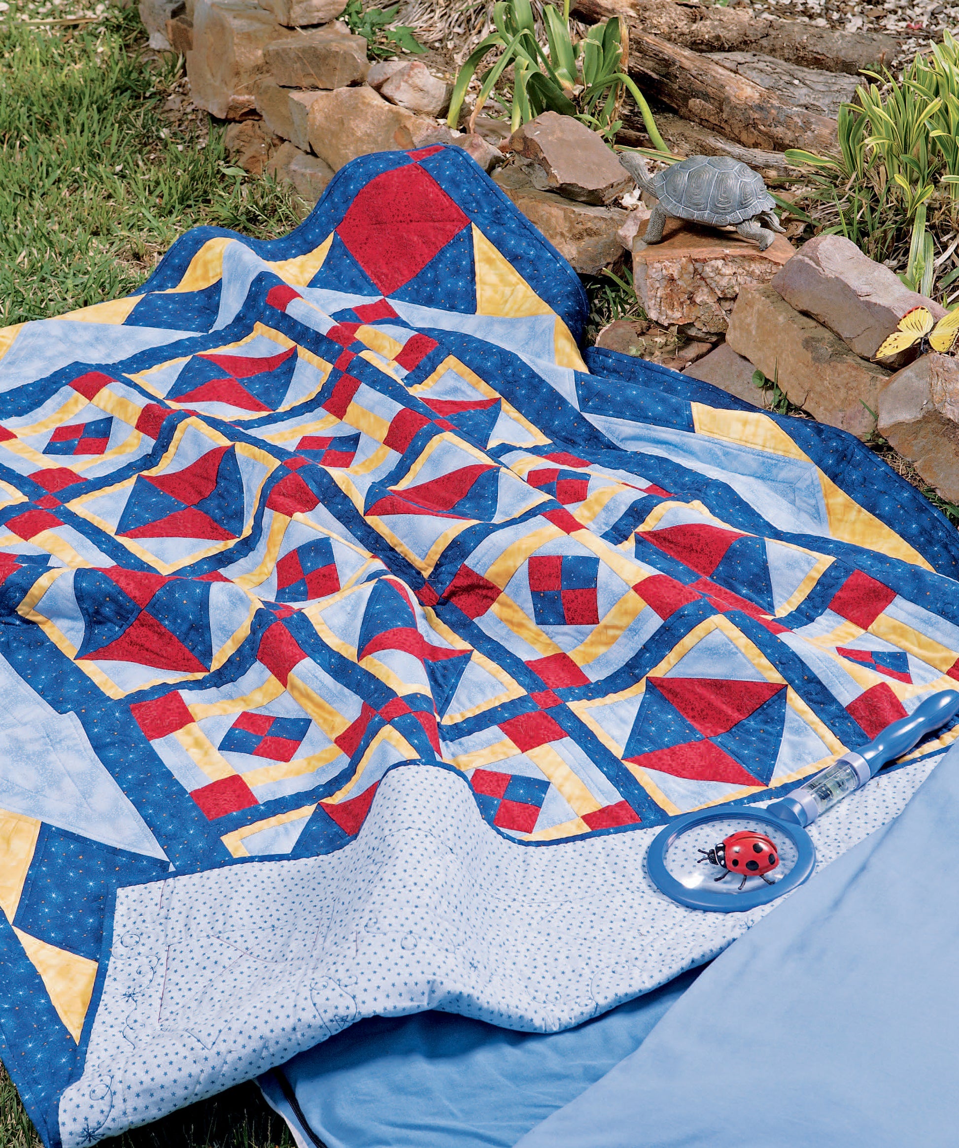 The Quilts for Kids—From Crib to College quilt by Leisure Arts, featuring red, blue, and yellow geometric patterns, is spread on grass beside a stone border with a toy ladybug on top and a gray turtle nearby.