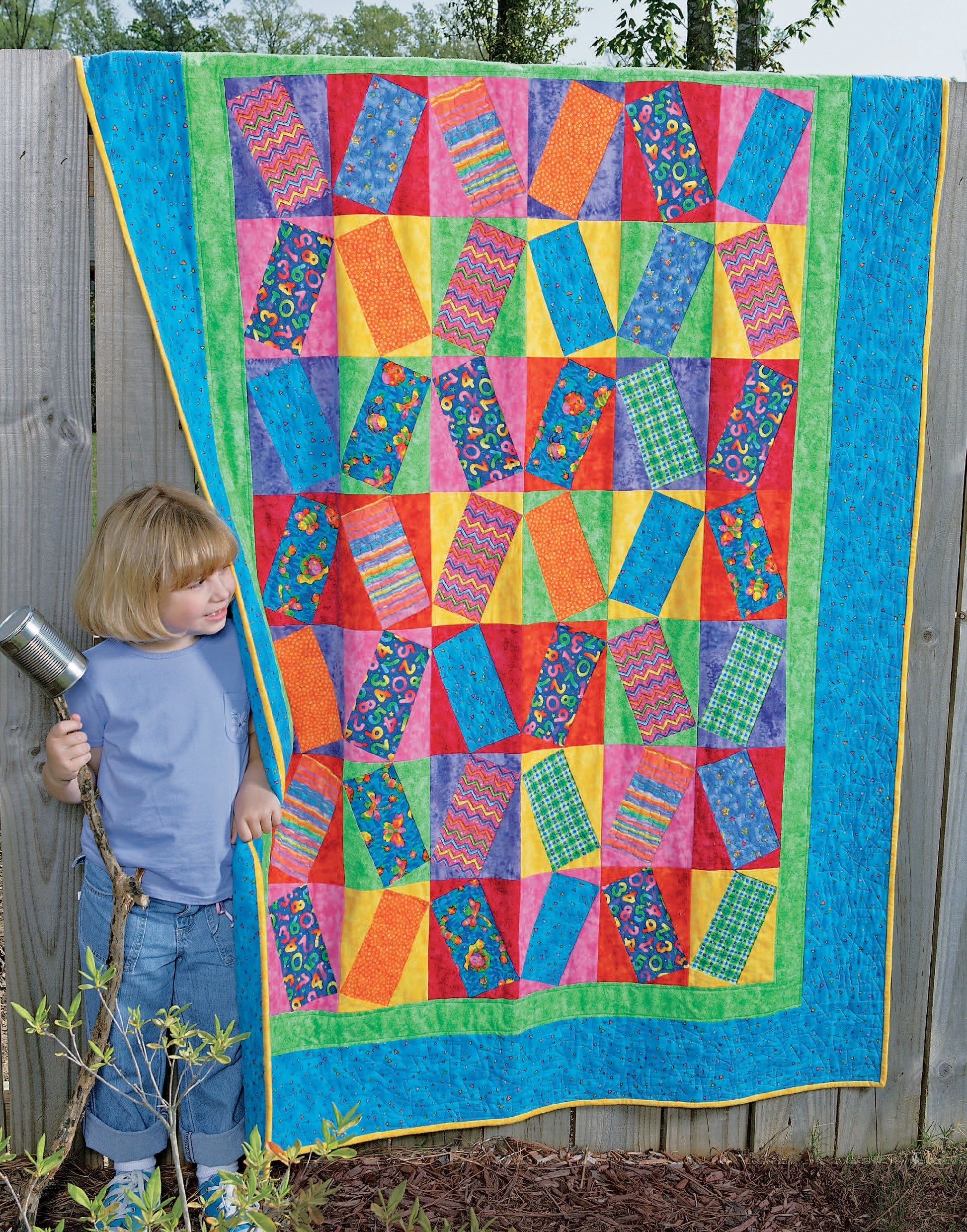A young child stands beside a vibrant patchwork quilt from "Quilts for Kids—From Crib to College" by Leisure Arts, featuring bold geometric patterns and a green-blue border, hanging on a wooden fence outdoors.