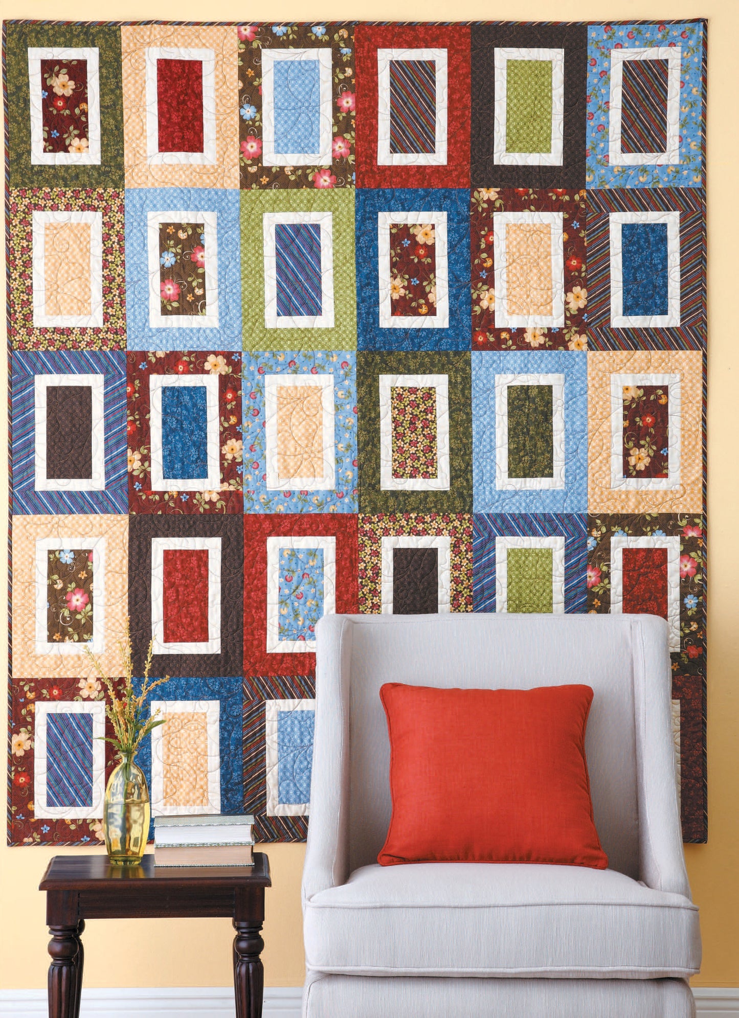 The Leisure Arts “Quilts for Kids—From Crib to College” patchwork quilt with rectangular patterns hangs on a yellow wall behind a light gray armchair with an orange cushion, beside a small side table holding a book, vase, and flowers.