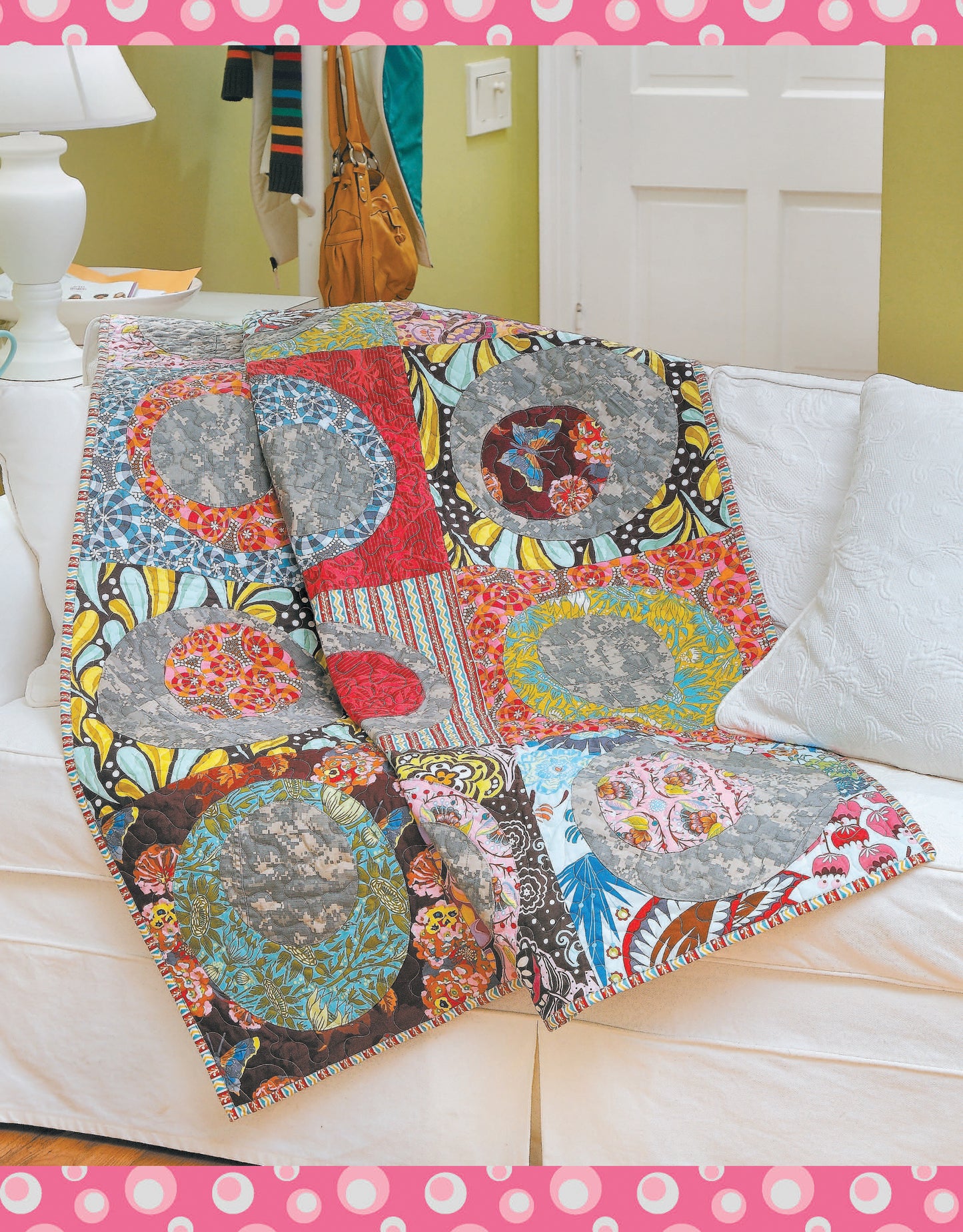 The Fun Colorful Quilts Digital Download by Leisure Arts features vibrant quilts with circular quilting, shown draped over a white couch in a cozy room with a green wall, white door, and bags by the door, framed by a pink polka-dot border.