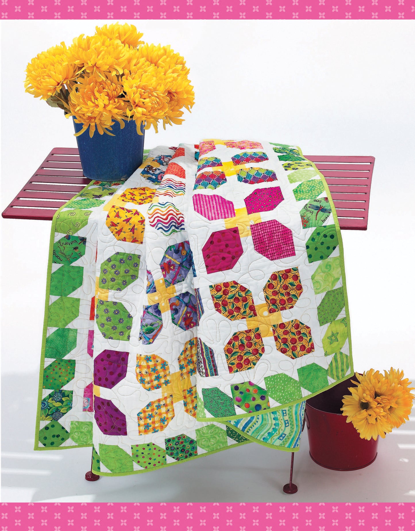 The Fun Colorful Quilts Digital Download by Leisure Arts features vibrant green, pink, and yellow quilting patterns draped over a red slatted table with a blue vase of yellow flowers on top and a red pail of blooms beside it.