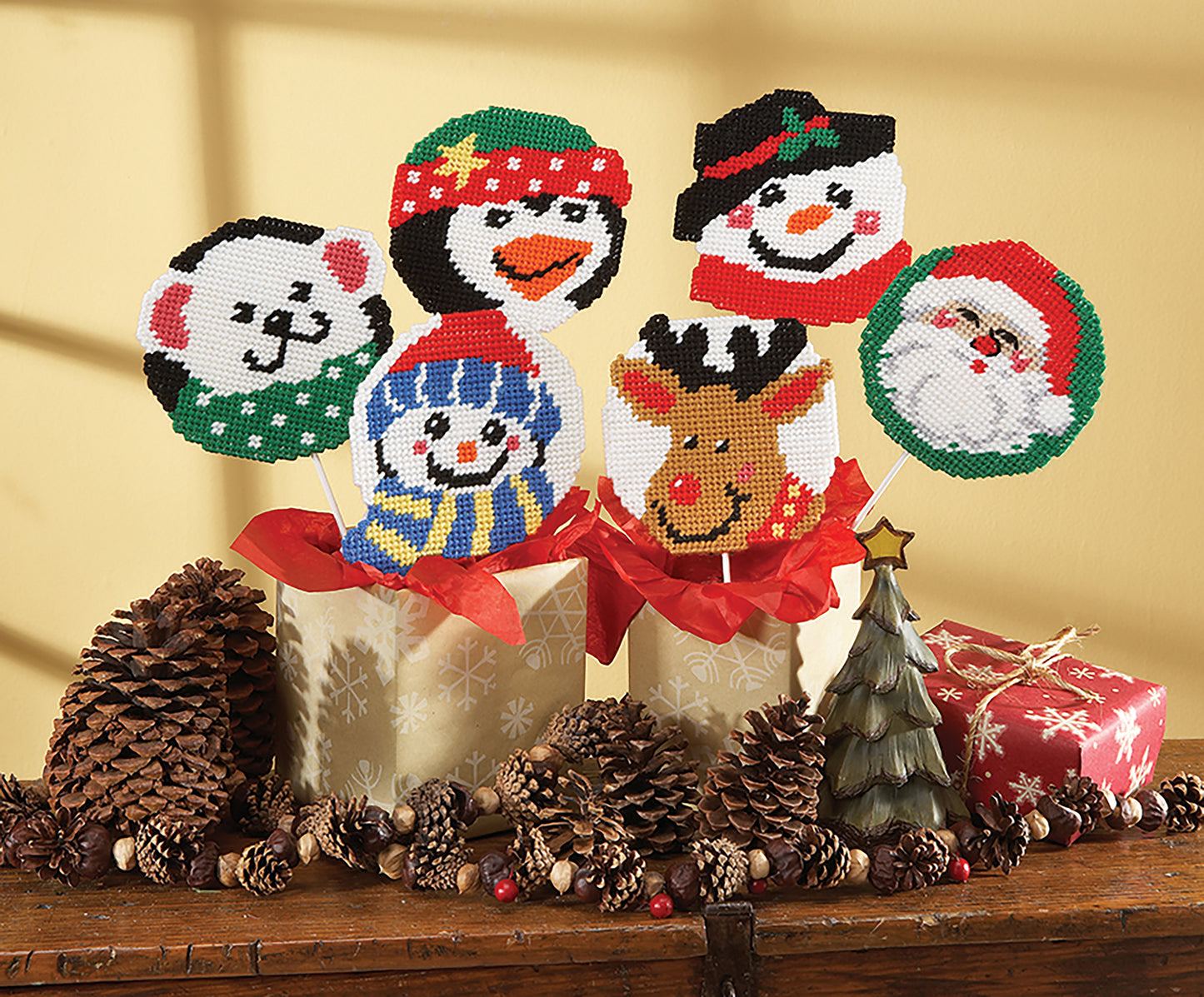 Six Coasters and Lollipop Covers by Leisure Arts, made with plastic canvas and featuring festive designs, are displayed in gift boxes with pinecones, a holiday ornament, and a wrapped present for charming seasonal accents.