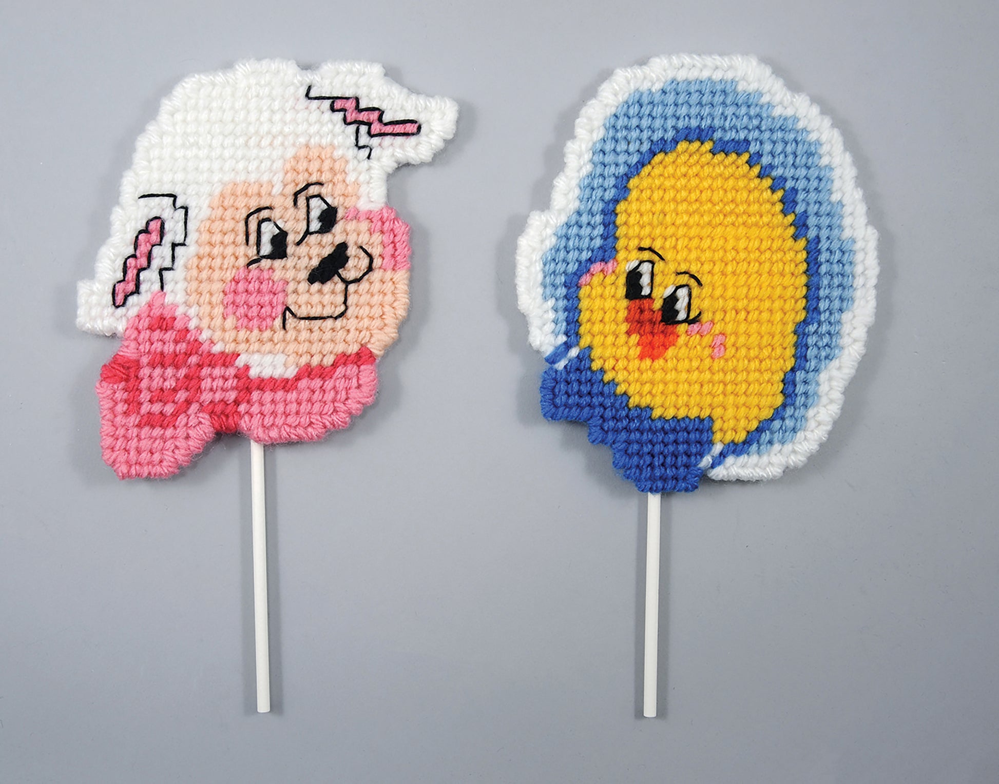 Leisure Arts Coasters and Lollipop Covers include two handmade plastic canvas covers: one with a cartoon lamb and pink bow, the other a smiling yellow chick with blue accents on white sticks—ideal festive gift accents.