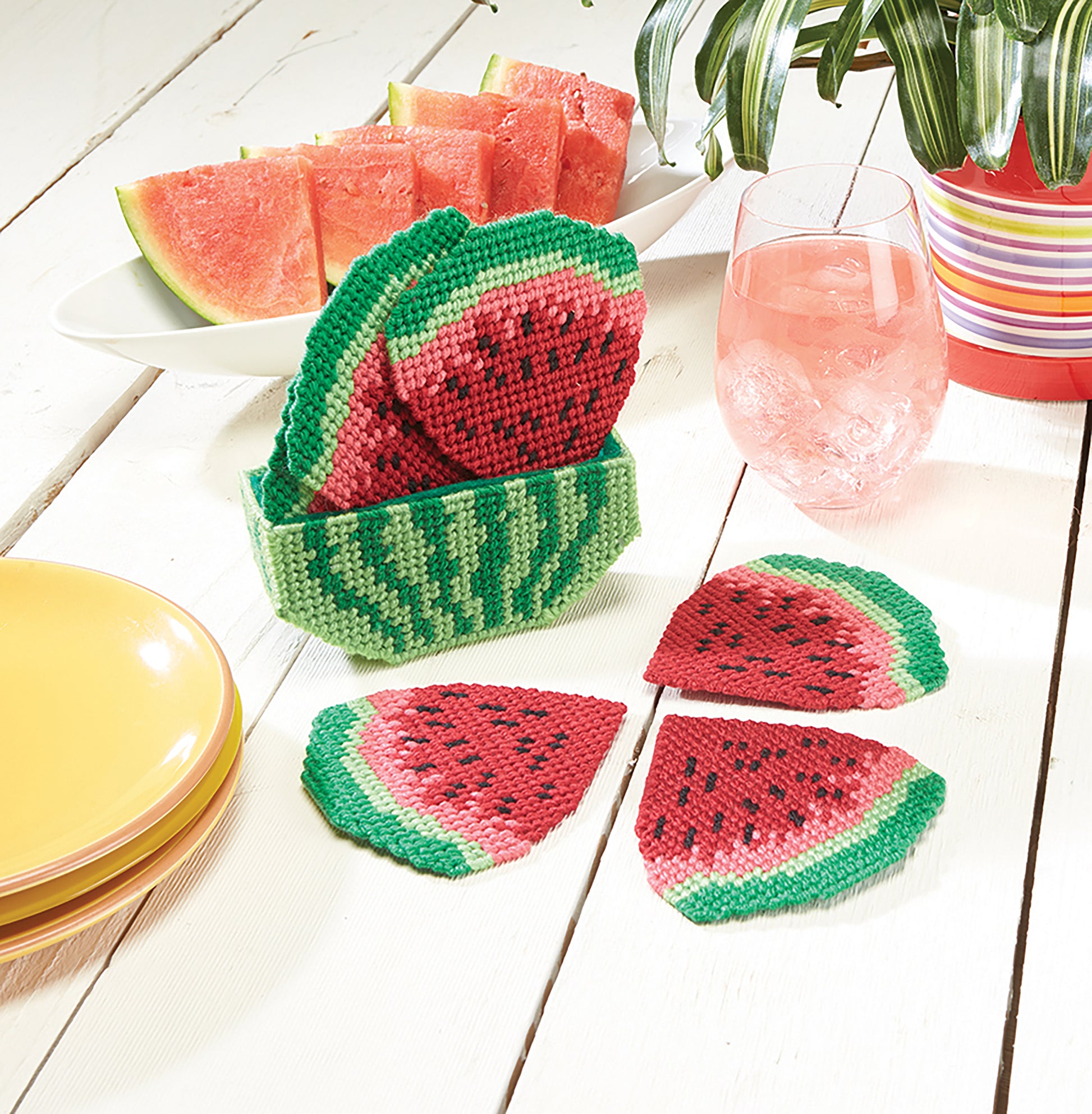 Leisure Arts Coasters and Lollipop Covers, featuring colorful watermelon slice designs, sit on a white table with plates, a pink drink, fresh watermelon slices, and a striped potted plant.