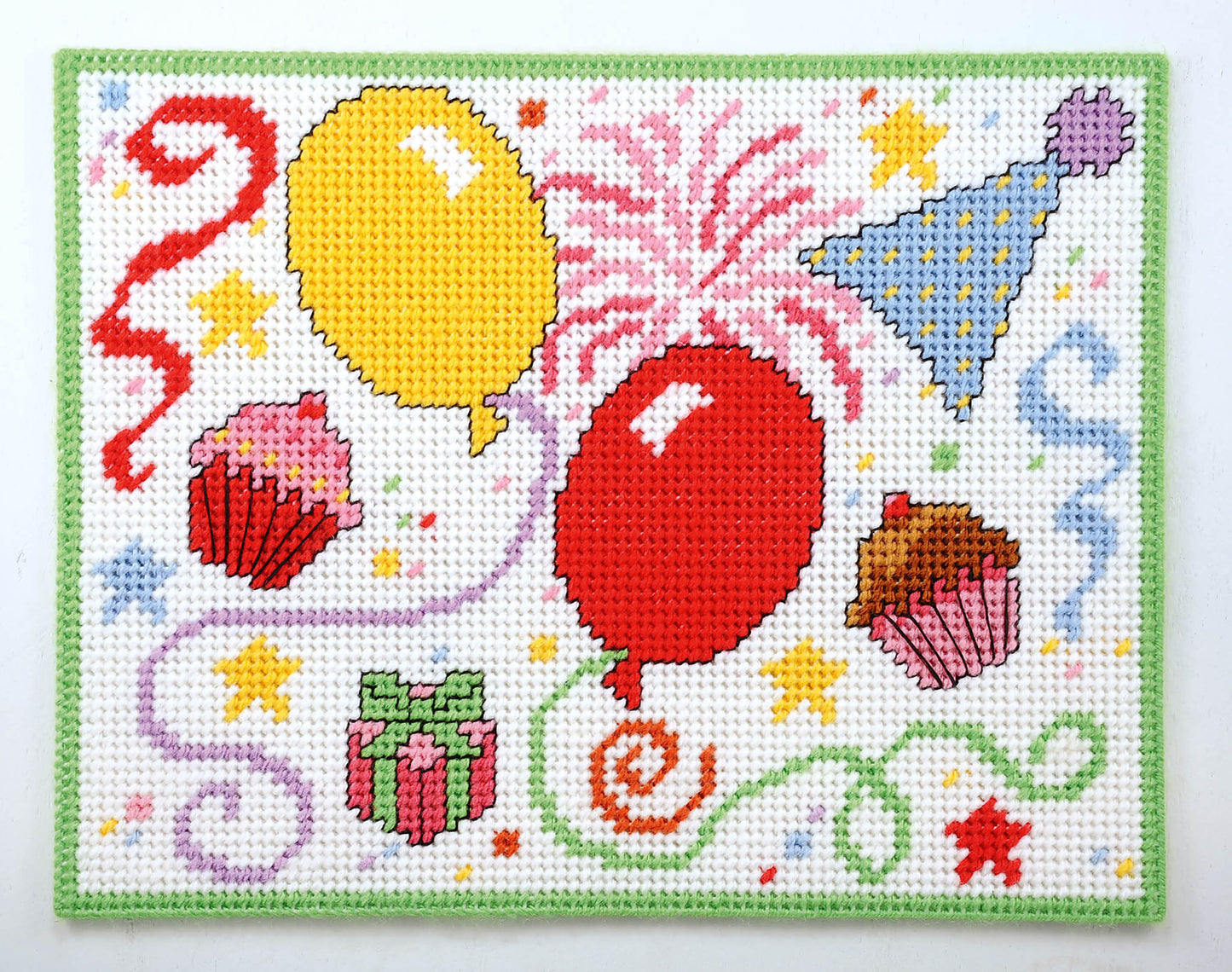 Party Place Mats by Leisure Arts feature a vibrant needlepoint design with balloons, cupcakes, party hat, present, streamers, fireworks, and stars on a white background with a green border—perfect for festive table settings.
