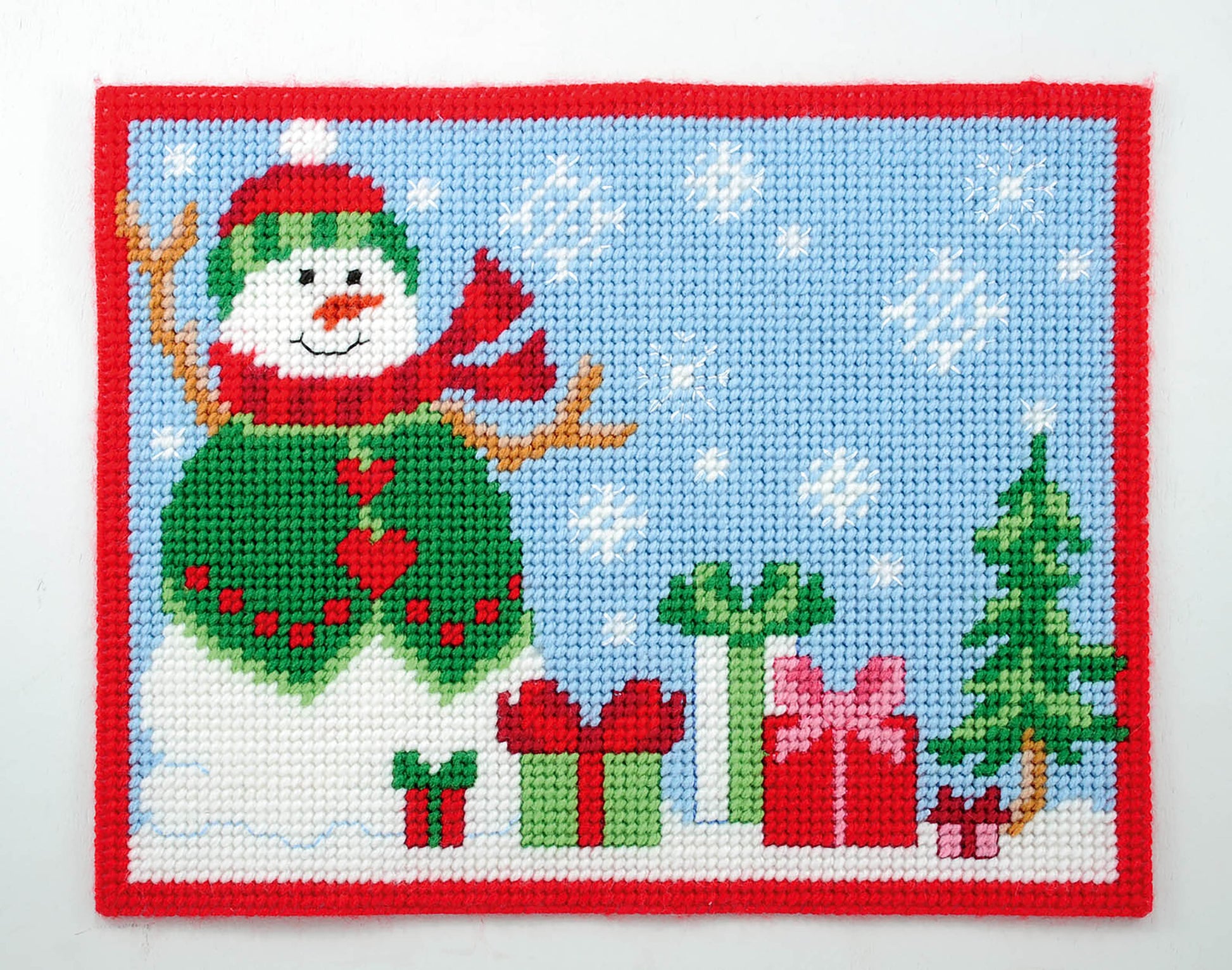 Create a festive table with the Leisure Arts Party Place Mats, featuring a cross-stitched snowman in a green sweater and red hat, surrounded by presents, a small tree, and snowflakes on light blue with a red border.