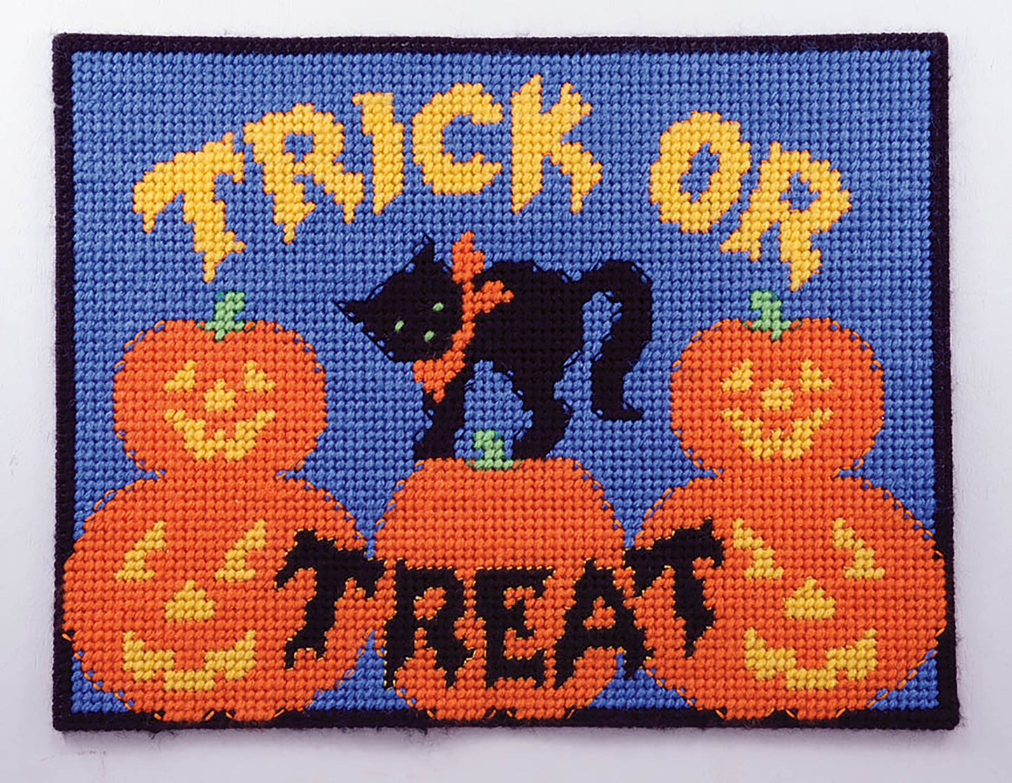 Leisure Arts Party Place Mats: A needlepoint design for plastic canvas featuring “Trick or Treat” in yellow, a black cat with an orange bow, and four smiling jack-o-lanterns—perfect for festive table settings on blue.