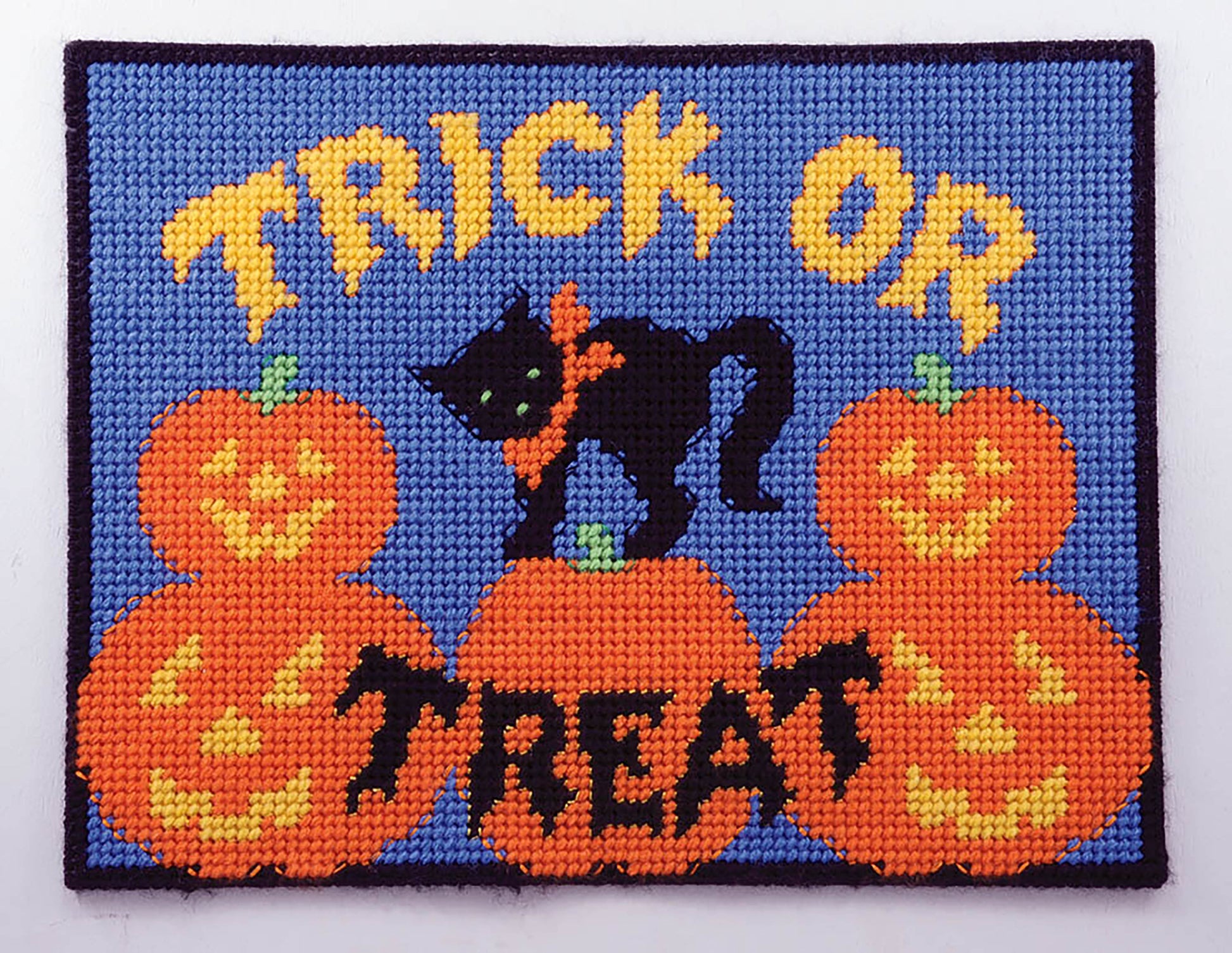 Leisure Arts Party Place Mats: A needlepoint design for plastic canvas featuring “Trick or Treat” in yellow, a black cat with an orange bow, and four smiling jack-o-lanterns—perfect for festive table settings on blue.