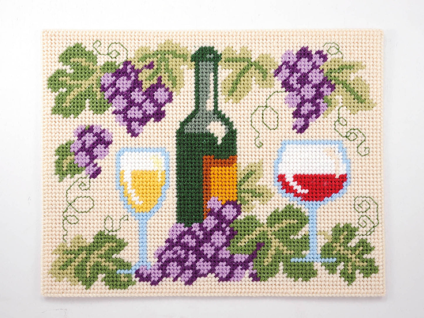 The Leisure Arts Party Place Mats cross-stitch pattern features a wine bottle, red and white wine glasses, grapes, and grape leaves on cream—ideal for festive table settings or pairing with plastic canvas place mats.