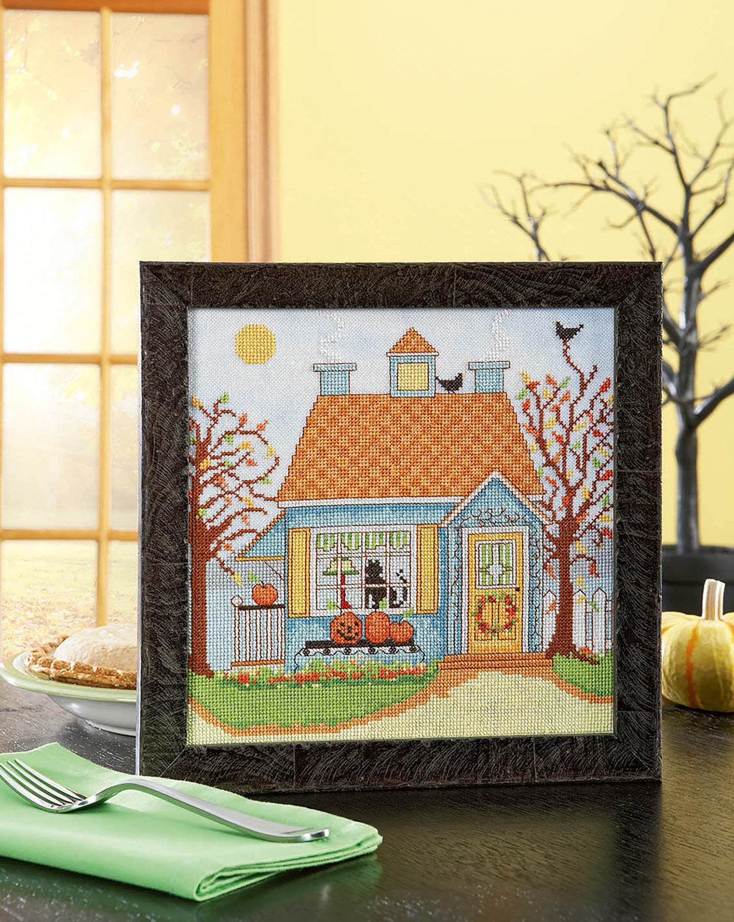 The "Seasonal Cottages Cross Stitch" by Leisure Arts features a blue house on hand-dyed fabric, with pumpkins in the yard, autumn trees, a black cat on the roof, plus window details and seasonal accents like pie and a green napkin.