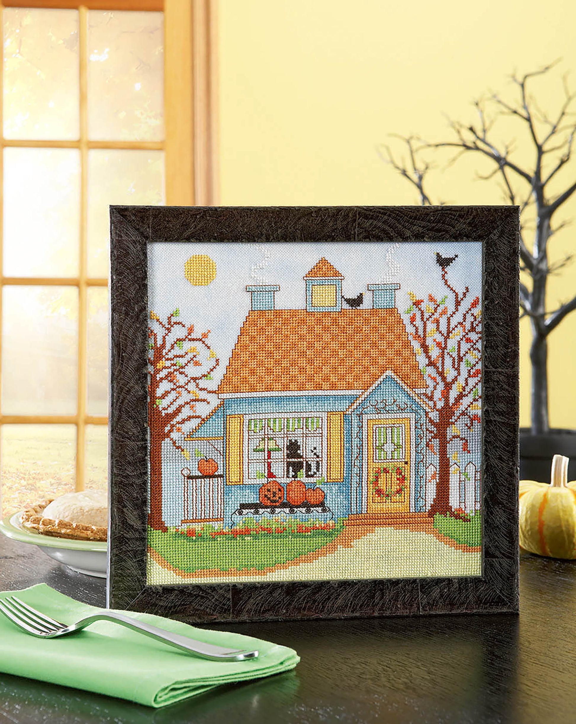The "Seasonal Cottages Cross Stitch" by Leisure Arts features a blue house on hand-dyed fabric, with pumpkins in the yard, autumn trees, a black cat on the roof, plus window details and seasonal accents like pie and a green napkin.