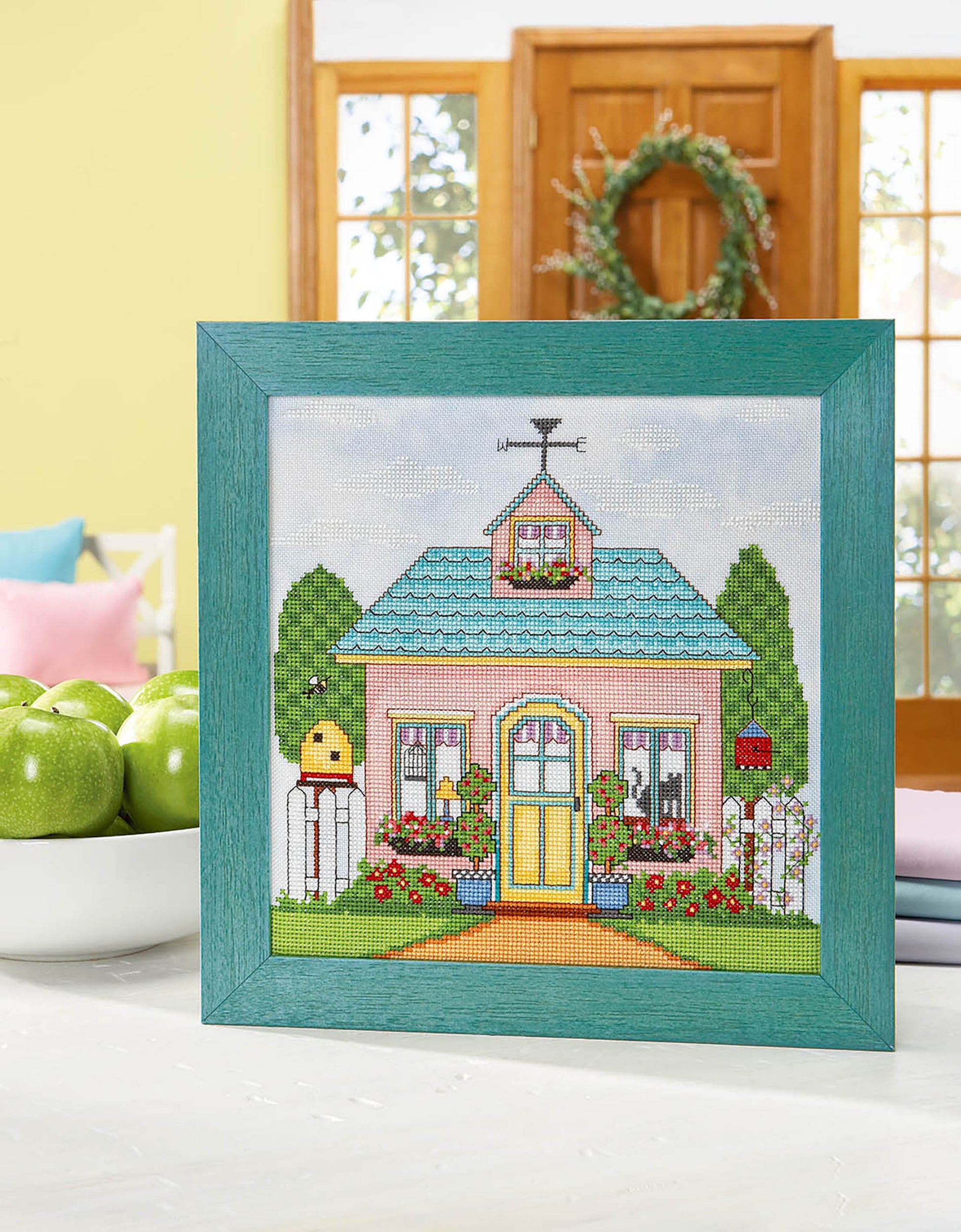 The Leisure Arts Seasonal Cottages Cross Stitch features a colorful house with a blue roof, yellow door, and garden on hand-dyed fabric, framed and displayed with green apples in a bright, cozy room.