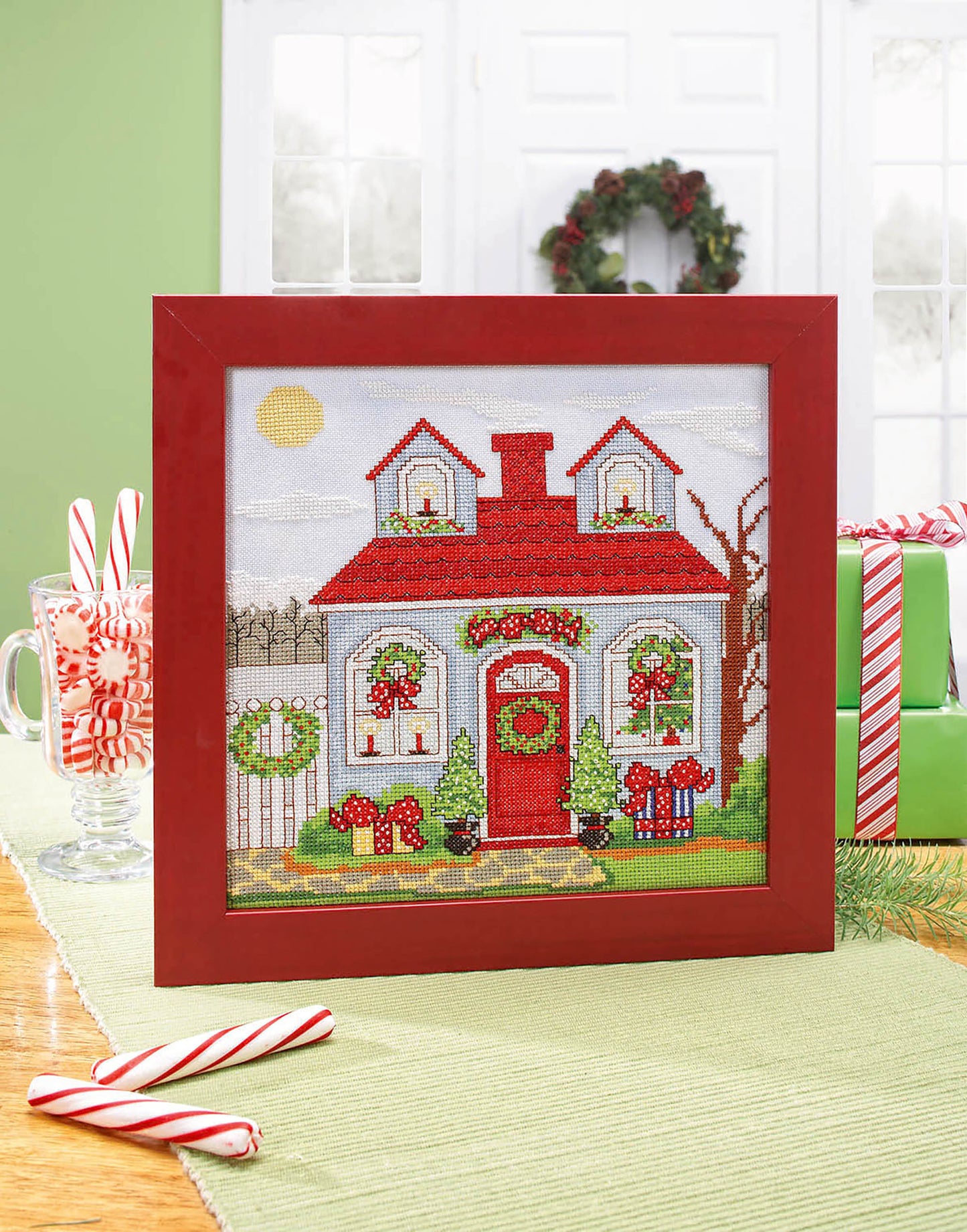 The Leisure Arts Seasonal Cottages Cross Stitch features a festive house with a red roof, green wreaths, and holiday decor on hand-dyed fabric, displayed with candy canes, wrapped gifts, and a white door adorned with a holiday wreath.