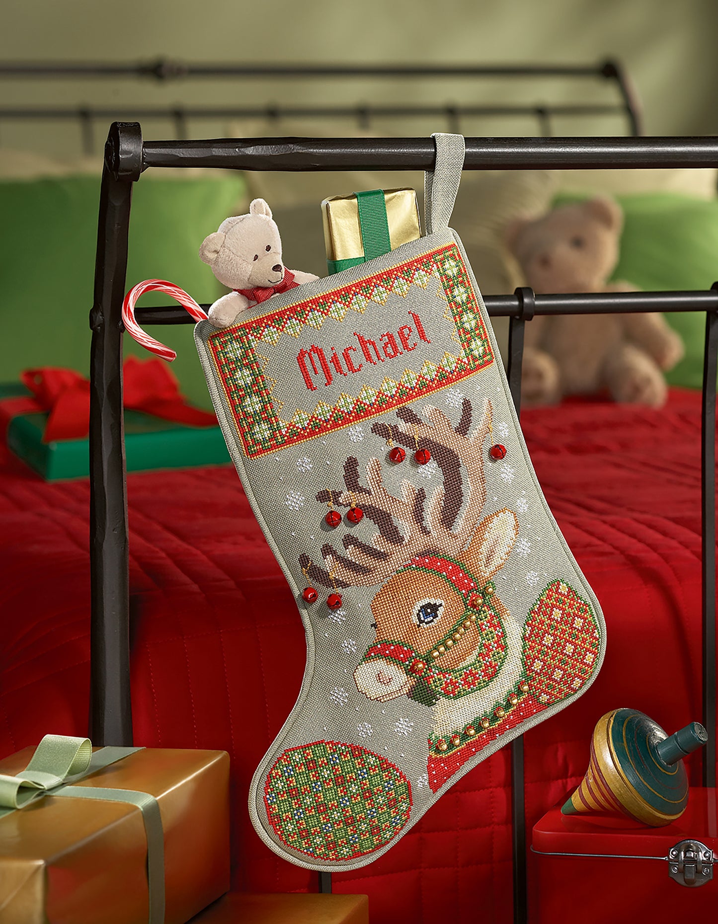 A Leisure Arts Christmas Stocking hangs on a bed frame, personalized with "Michael" and a reindeer design. Perfect for beginners, it holds a teddy bear, gold gift, and candy cane, with extra gifts displayed on the bed.