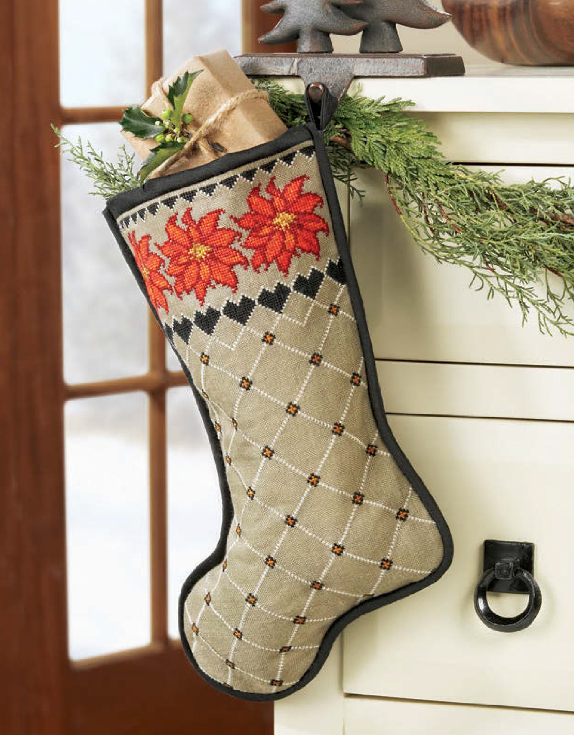 The Leisure Arts Christmas Stockings feature a diamond pattern with red poinsettias and hang from a white dresser with greenery. Rolled brown paper and gold ribbon are inside, making it easy to add a personalized alphabet—even for beginners.