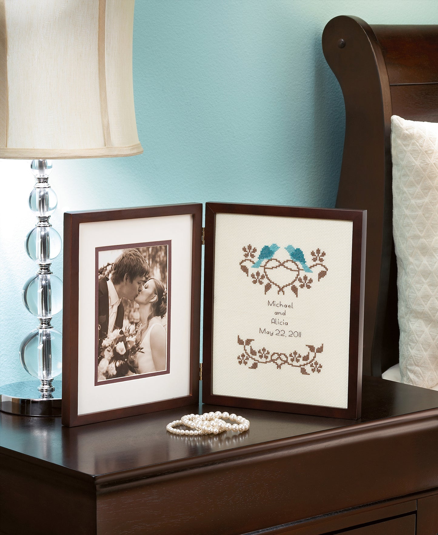 A hinged double frame on a nightstand displays a sepia wedding photo and a Leisure Arts "Wedding Samplers—Choose From Modern or Traditional Style!" cross stitch, handmade with blue birds, a heart, and “Michael and Alicia, May 22, 2011.”.