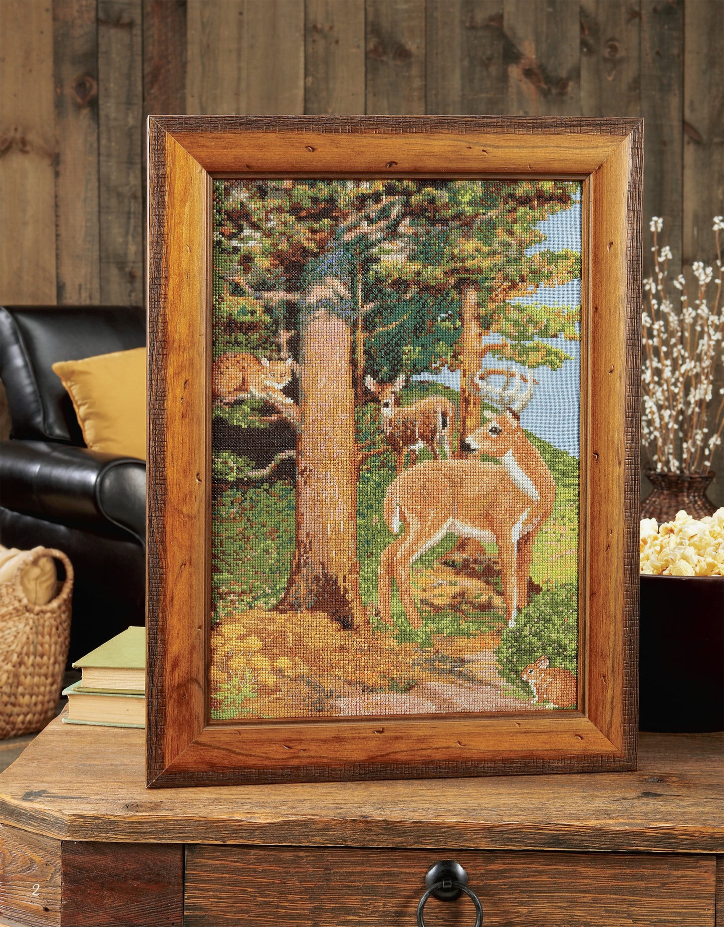 Leisure Arts’ Cross Stitch Wildlife Portraits features three deer in a forest, framed and displayed on a wooden table—an ideal rustic home decor piece for nature lovers.