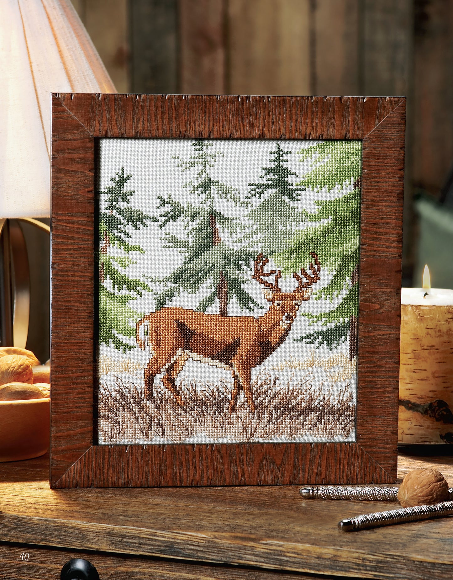 Leisure Arts’ Cross Stitch Wildlife Portraits showcases a majestic stag in grass with evergreens, framed and displayed on a rustic wooden surface—ideal home decor beside a lamp, candle, and small accents.