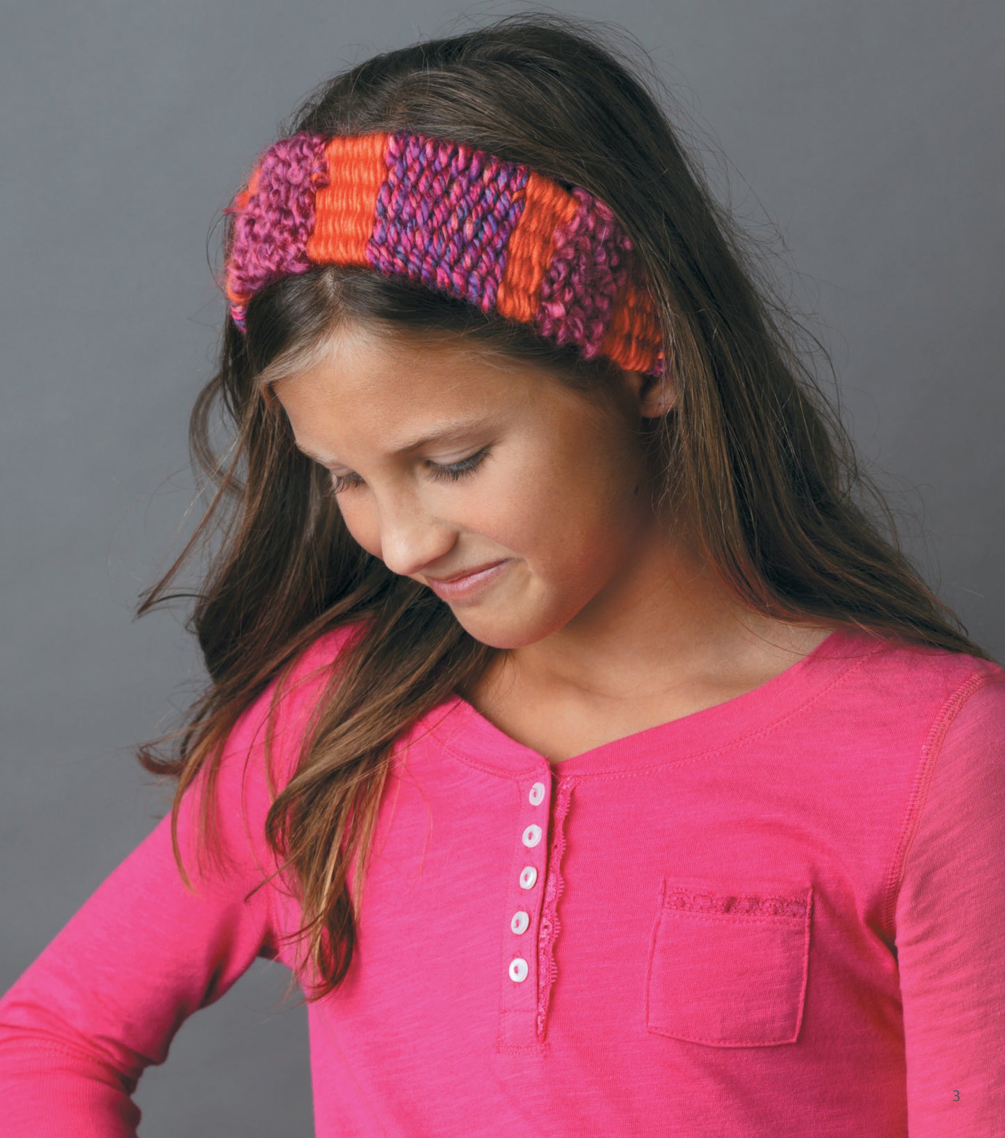 A young girl in a pink long-sleeve henley smiles softly, wearing a striped beginner headband she made with Leisure Arts’ The Dream Weaver—A Simple Way to Weave Anything You Can Imagine.