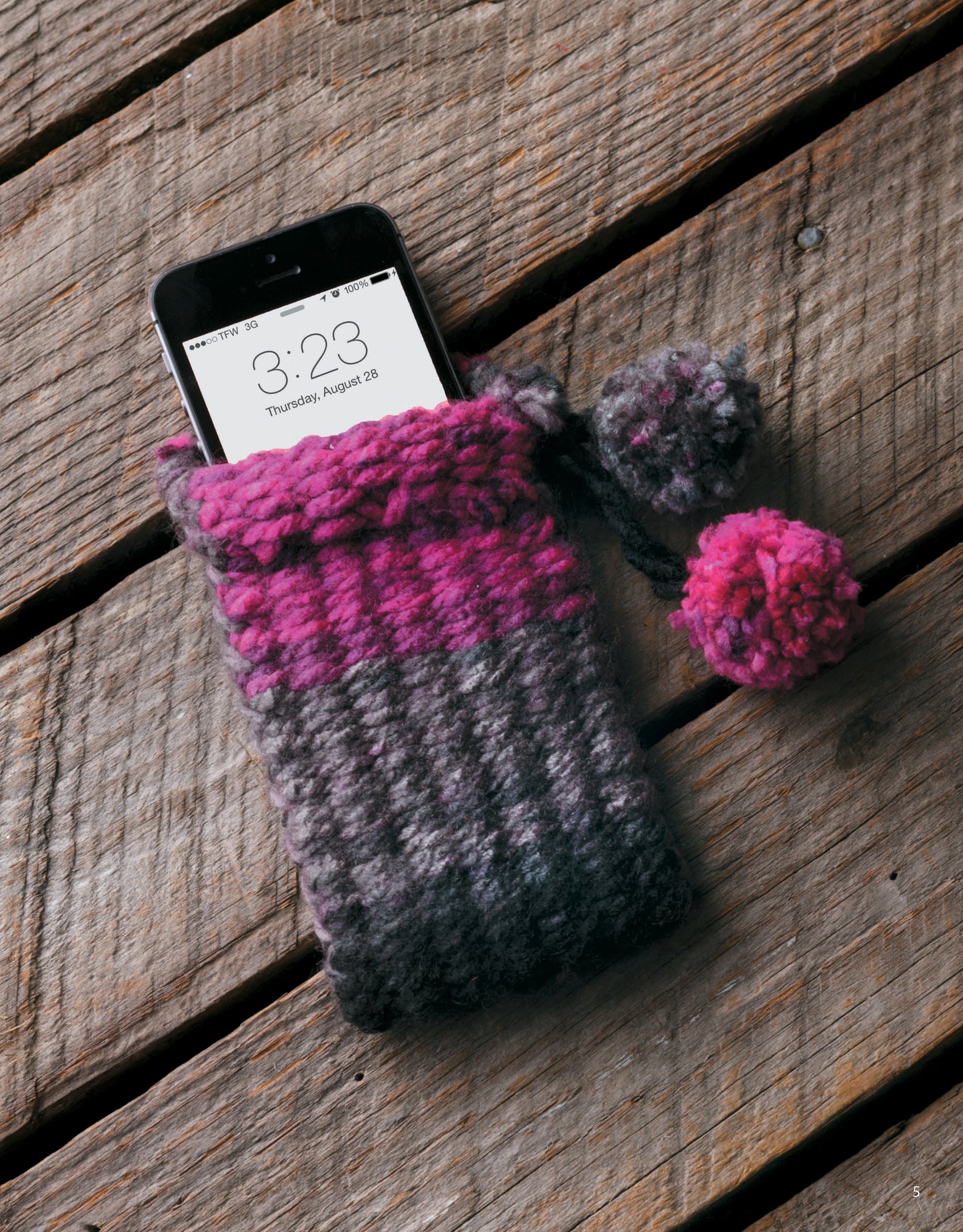 A smartphone sits in a gray and pink knitted pouch with pom-poms, crafted using The Dream Weaver by Leisure Arts, on a wooden surface. The phone displays the time as 3:23 and the date as Thursday, August 28.