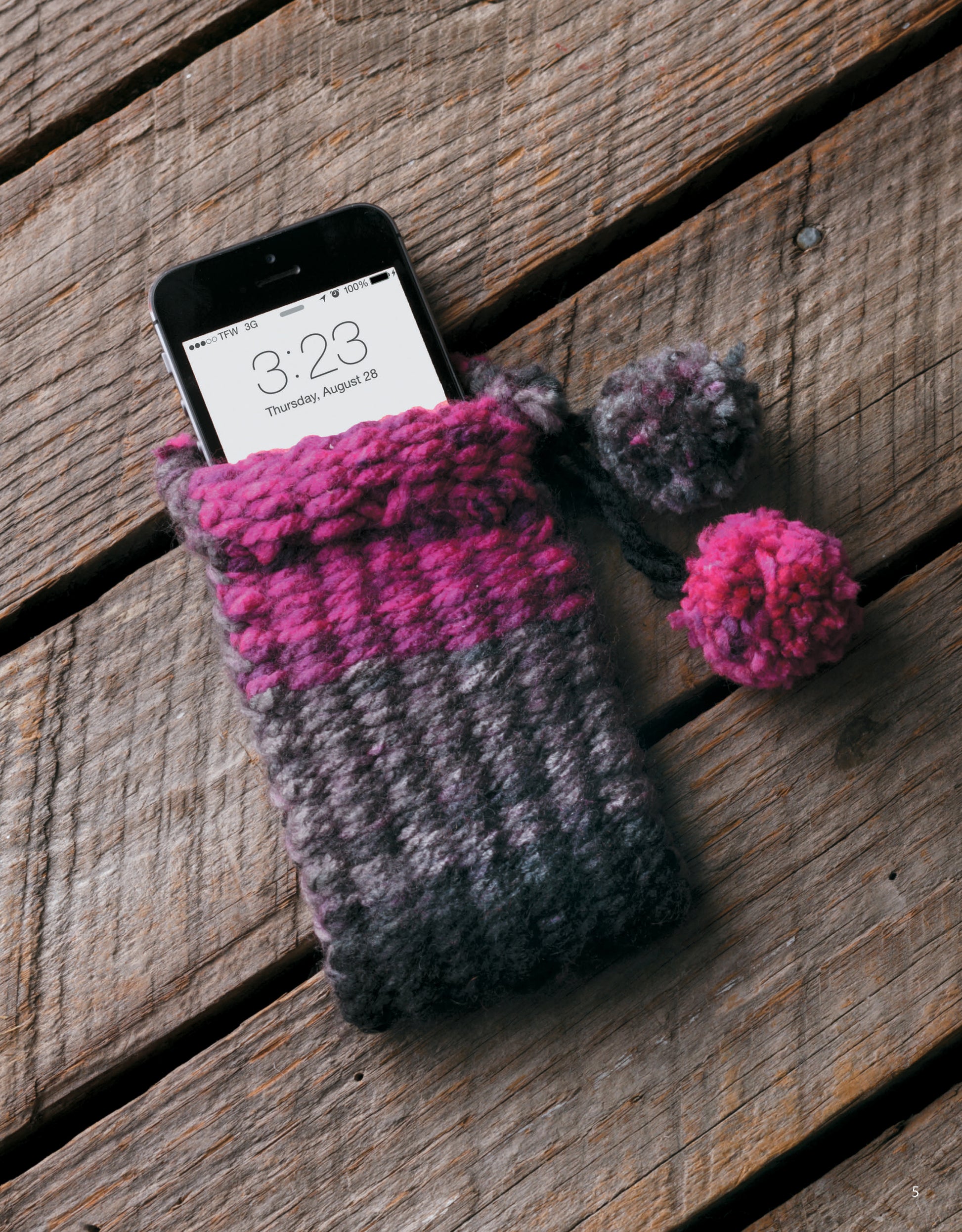 A smartphone sits in a gray and pink knitted pouch with pom-poms, crafted using The Dream Weaver by Leisure Arts, on a wooden surface. The phone displays the time as 3:23 and the date as Thursday, August 28.