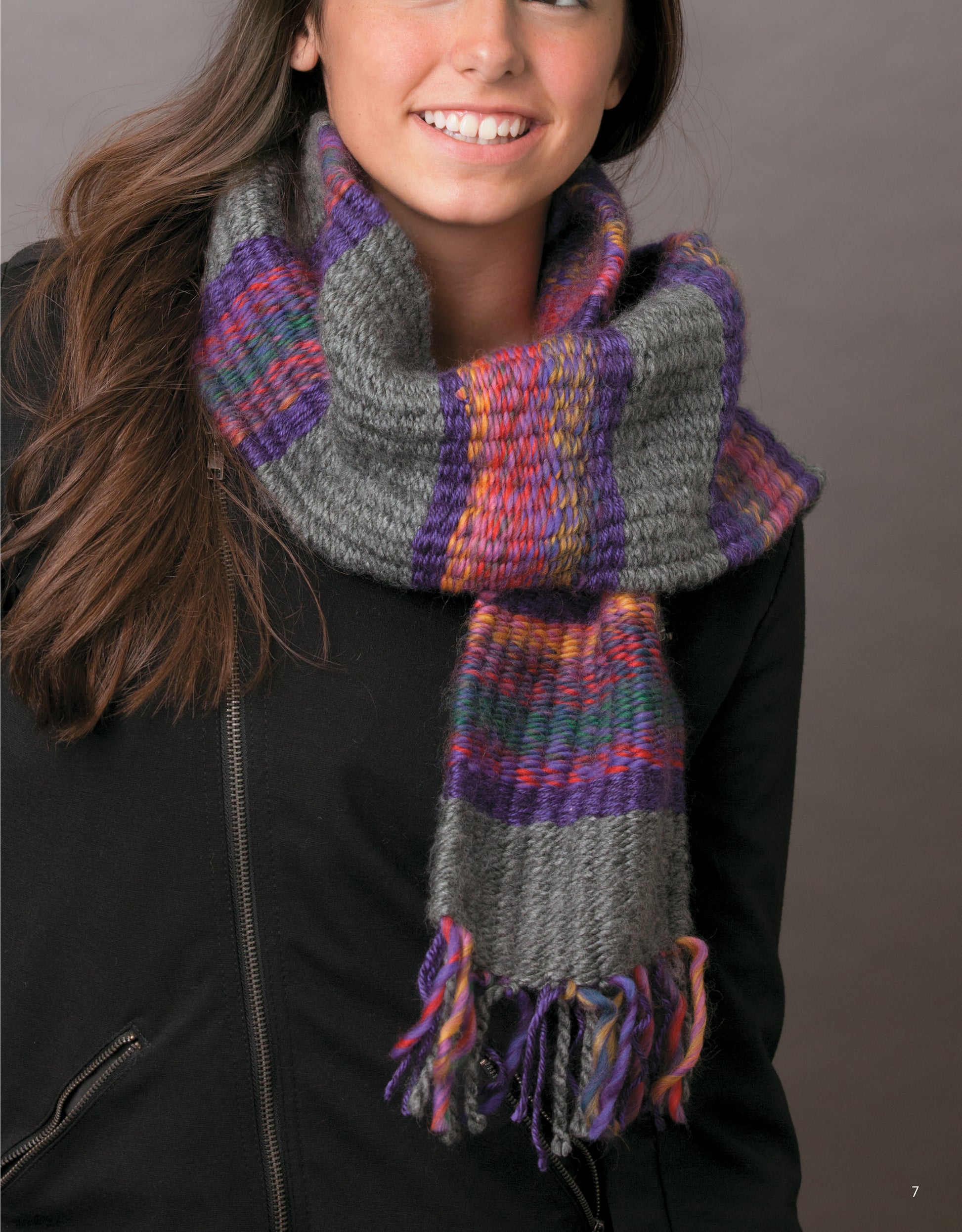 A person models a thick, fringed scarf with gray, purple, orange, and multicolor stripes—woven using Leisure Arts’ The Dream Weaver: A Simple Way to Weave Anything You Can Imagine—alongside a black jacket against a plain backdrop.