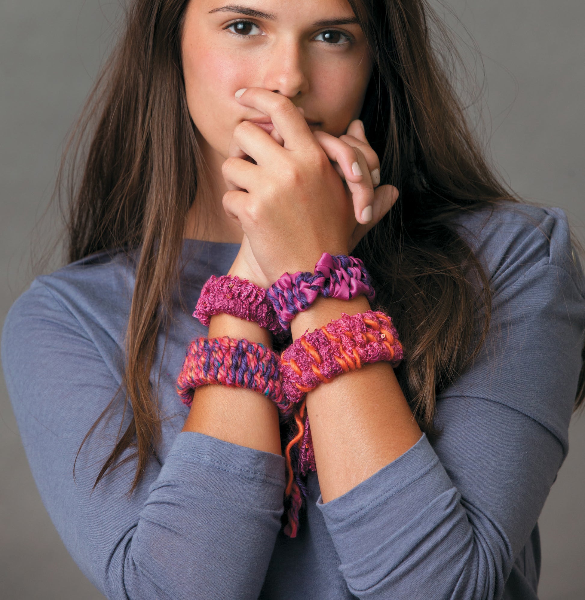 A person with long brown hair wears a gray long-sleeve shirt and displays several colorful bracelets made using The Dream Weaver by Leisure Arts, holding their hands near their face against a plain background.