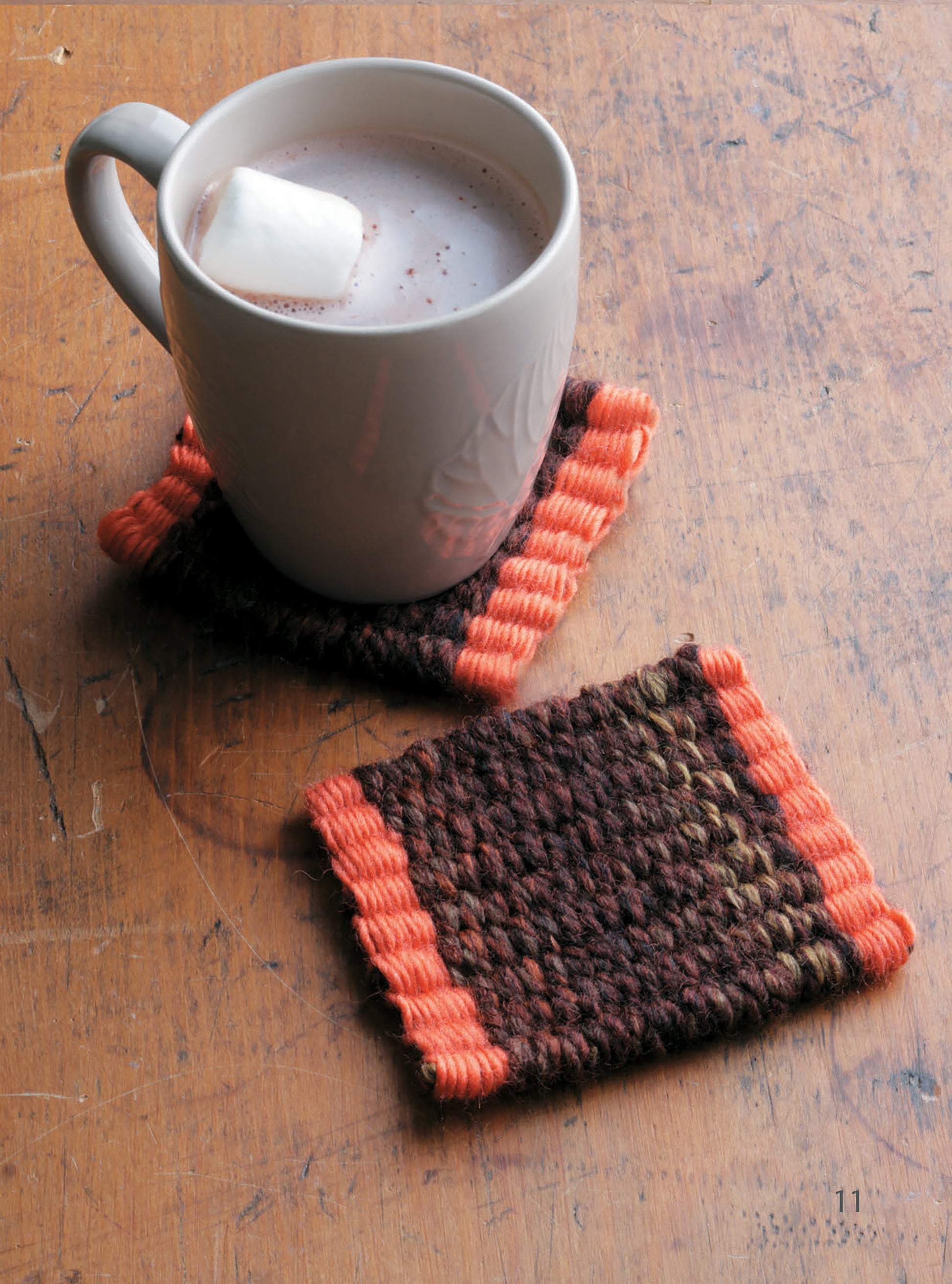 A white mug of hot cocoa with a marshmallow sits on a brown and orange coaster, handmade with The Dream Weaver by Leisure Arts. Another matching coaster rests nearby atop a worn wooden surface.