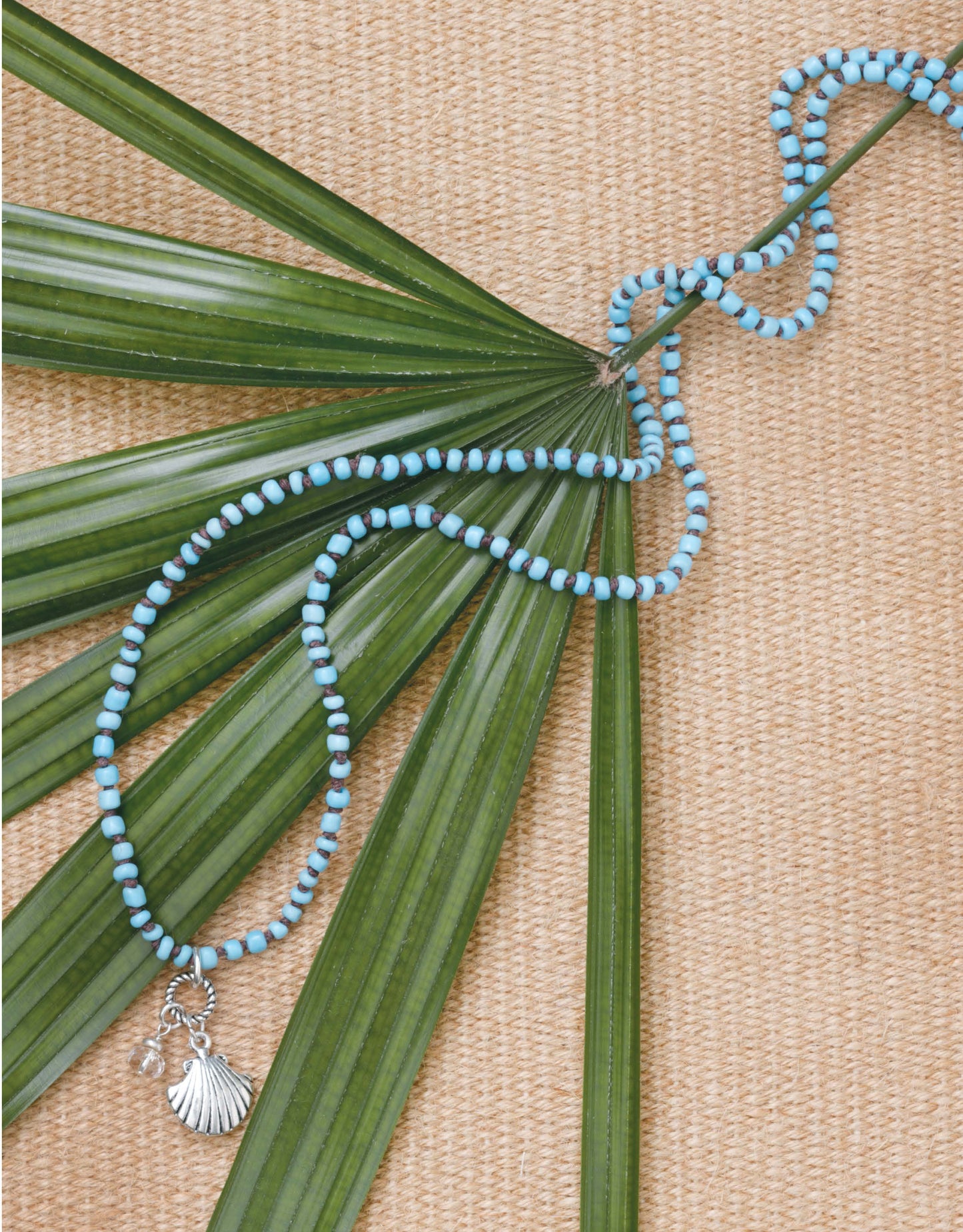Beach Chic Jewelry by Leisure Arts features alternating blue and white beads with a silver shell pendant, displayed on green palm leaves over a woven beige mat—ideal for fans of ocean-inspired accessories.