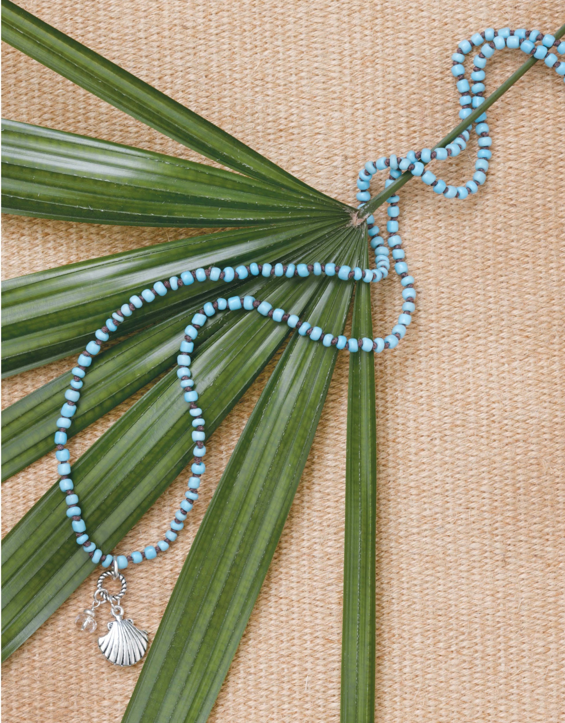 Beach Chic Jewelry by Leisure Arts features alternating blue and white beads with a silver shell pendant, displayed on green palm leaves over a woven beige mat—ideal for fans of ocean-inspired accessories.