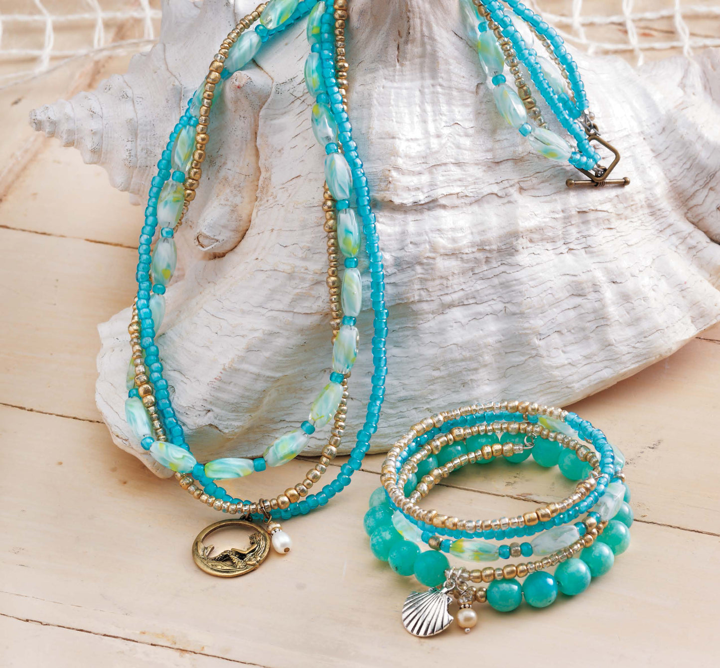 Beach Chic Jewelry by Leisure Arts features turquoise, gold, and pearl layered necklaces with a round pendant, shell charm, and matching bracelets, beautifully displayed on a white seashell over a light wooden surface.