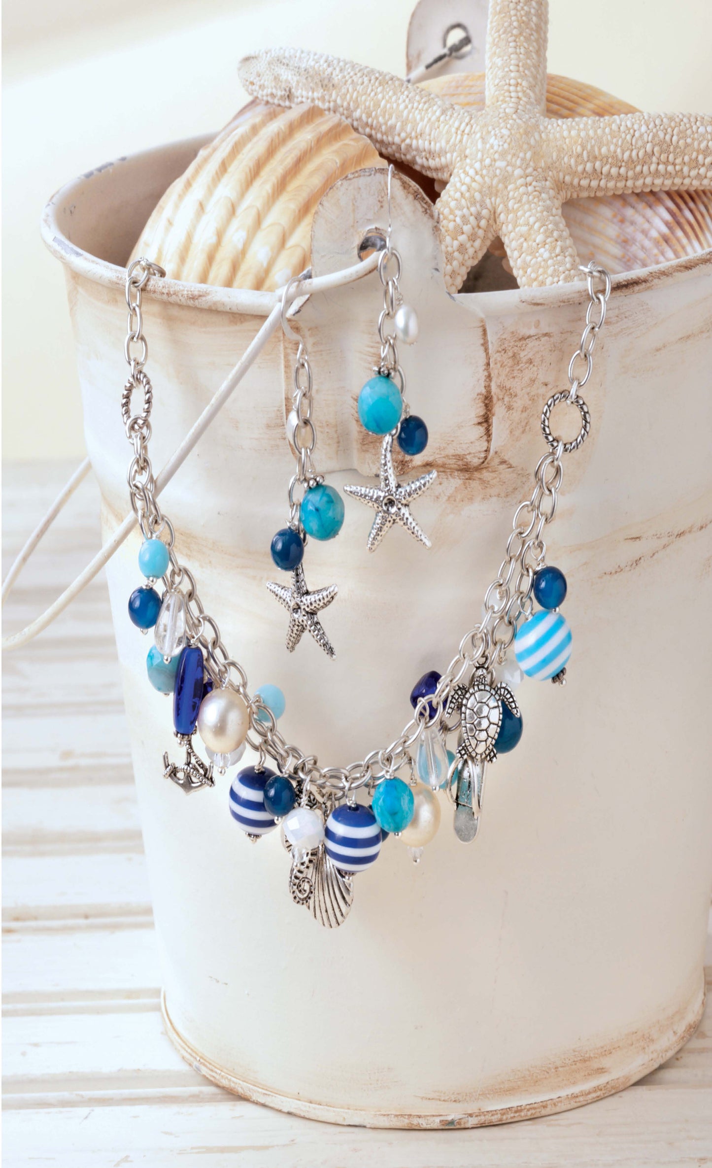 Beach Chic Jewelry by Leisure Arts—a coastal necklace and matching earrings with blue, white, and silver beads plus starfish, shell, and anchor charms—are displayed in a white bucket with seashells on a light wooden surface.