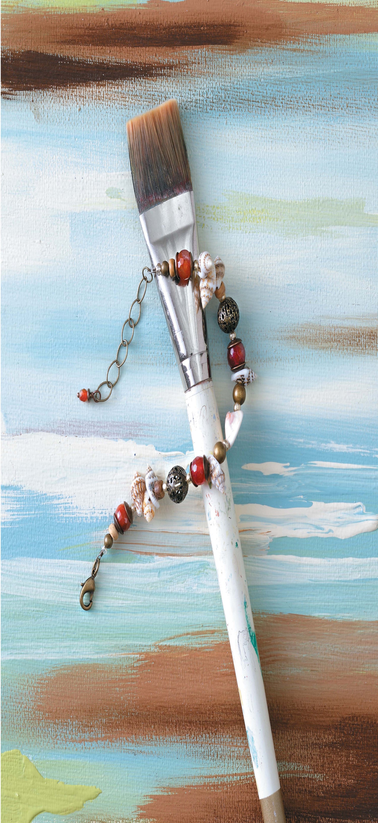 A Leisure Arts Beach Chic Jewelry bracelet, adorned with beads and shells, is displayed on a painted canvas with abstract blue, white, brown, and green strokes alongside a white paintbrush.