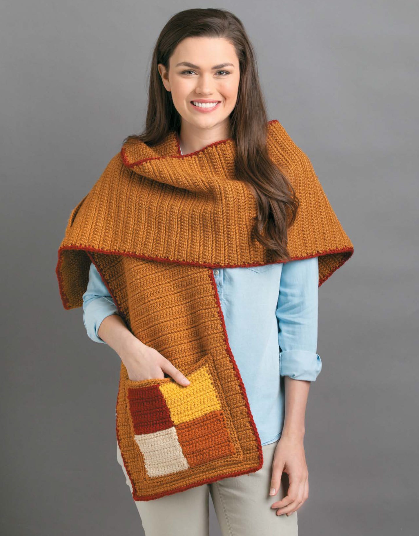 A woman in a light blue shirt smiles while modeling a large, rust-orange Pocket Shawls Digital Download by Leisure Arts, featuring yellow, rust, and cream patchwork pockets—a cozy accessory to showcase unique crochet shawl patterns.