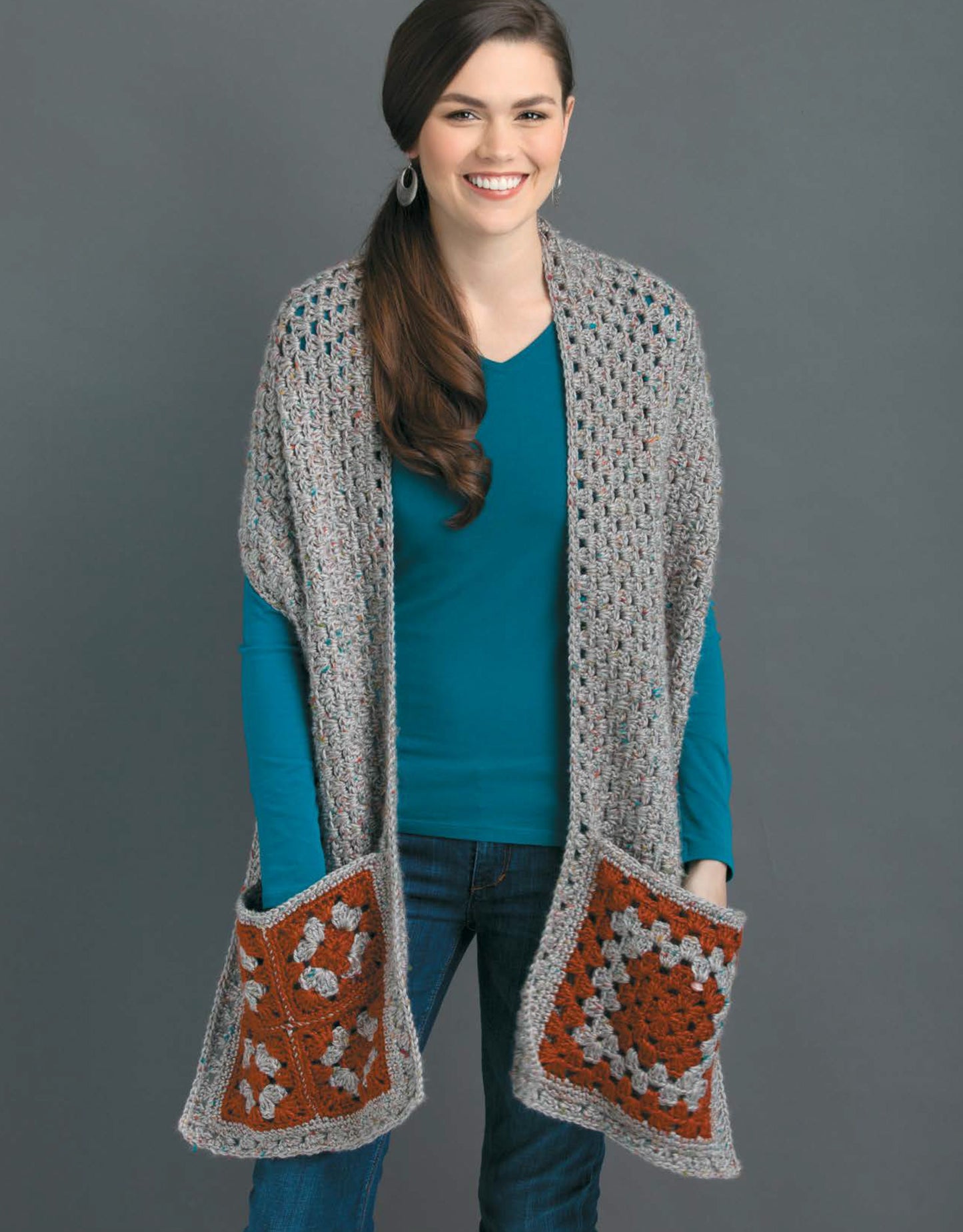 A smiling woman models the Pocket Shawls Digital Download by Leisure Arts, featuring a teal shirt and a gray crochet shawl with large orange and gray patterned pockets—ideal inspiration for your next crochet project.