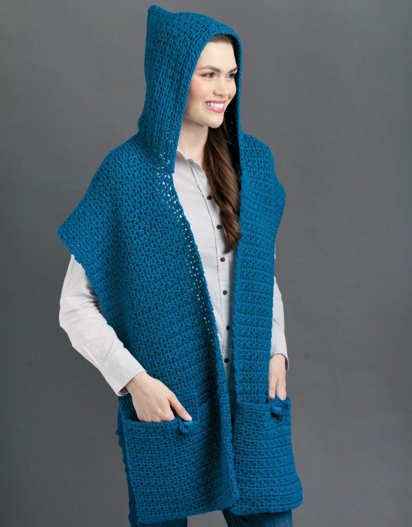 A woman smiles while modeling the Leisure Arts Pocket Shawls Digital Download—a crocheted bright blue shawl with a hood and pockets—over a light shirt and jeans, standing against a plain gray background.