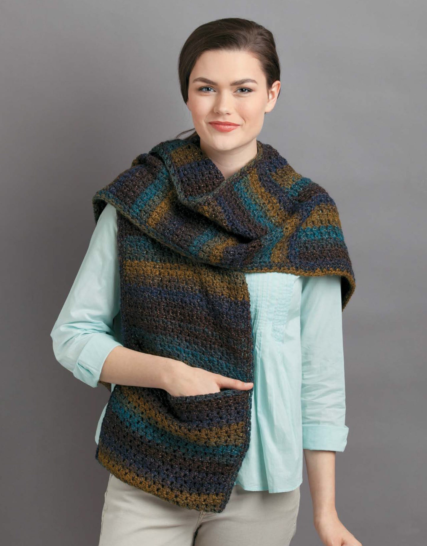 A model in a light blue shirt and beige pants showcases the Leisure Arts Pocket Shawls Digital Download, featuring a large striped crochet shawl in blue, green, brown, and gray against a plain gray background—great for adult or child projects.
