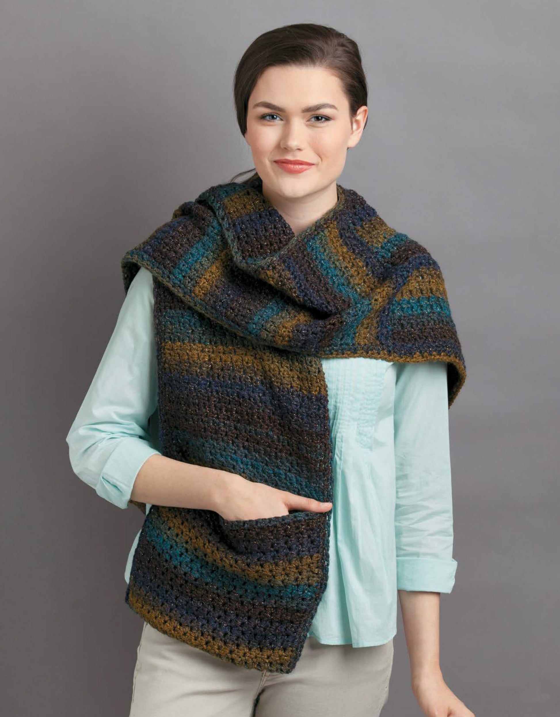 A model in a light blue shirt and beige pants showcases the Leisure Arts Pocket Shawls Digital Download, featuring a large striped crochet shawl in blue, green, brown, and gray against a plain gray background—great for adult or child projects.